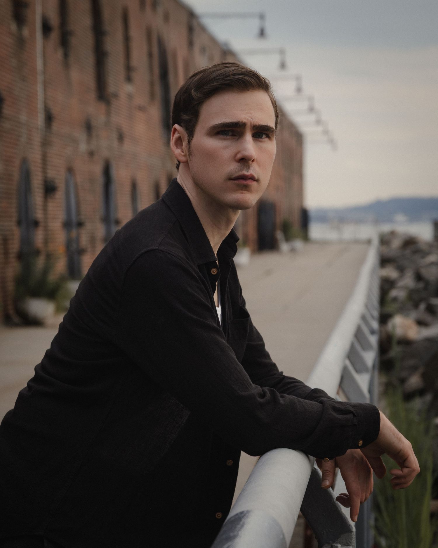 Dylan leans over a metal railing in a close waterfront portrait, with the brick warehouse and shoreline receding behind him.
