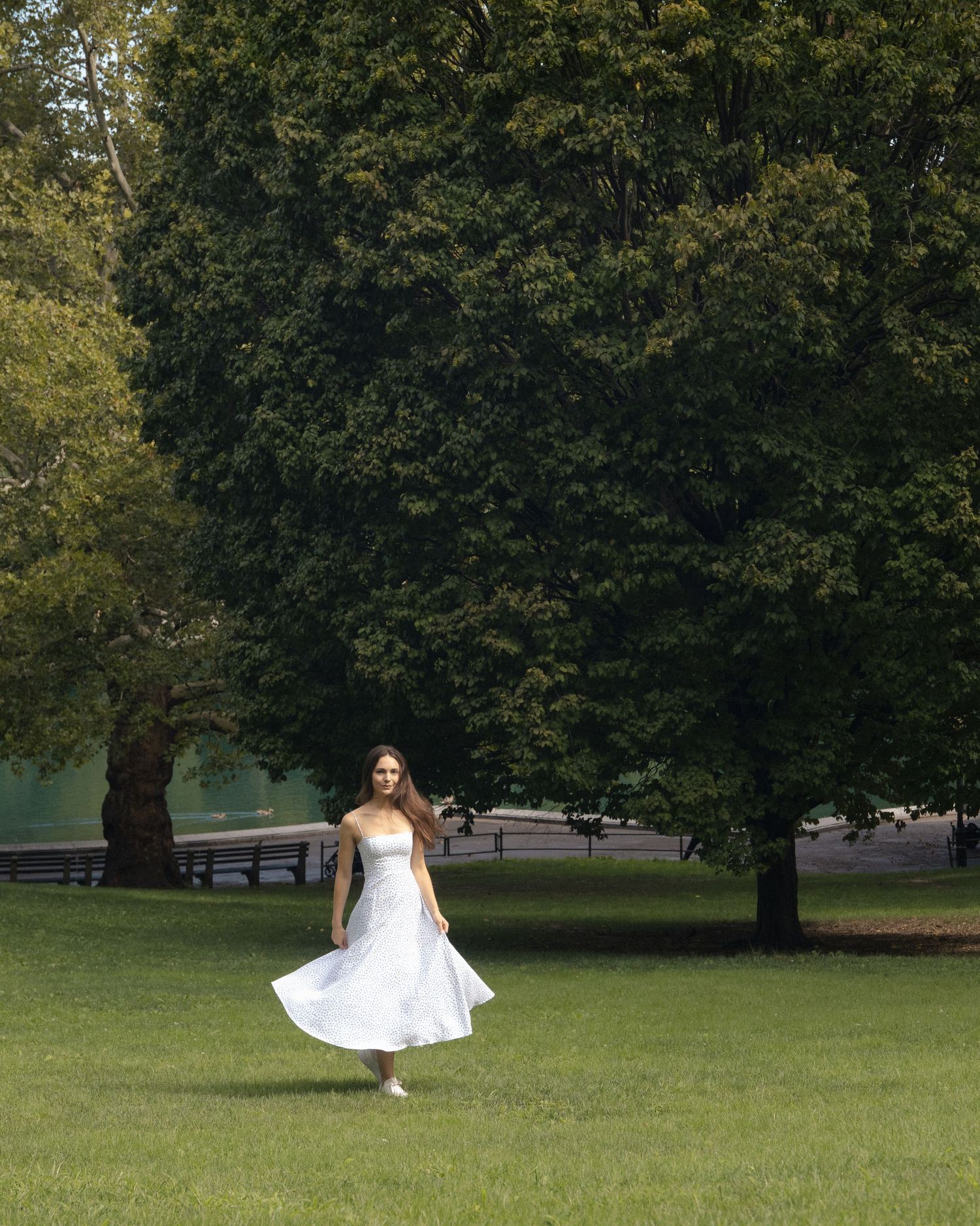 Woman walking across a green lawn, holding out her white floral skirt beneath the trees in Central Park.