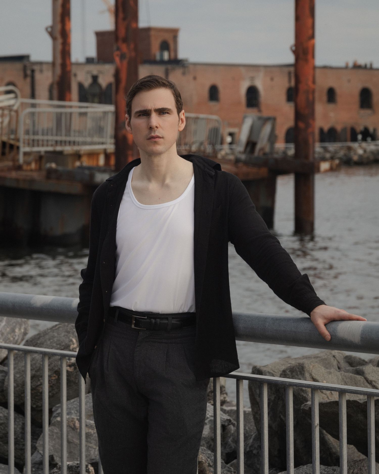 Dylan stands at a metal waterfront railing with weathered dock structures and an industrial brick building across the harbor in Red Hook