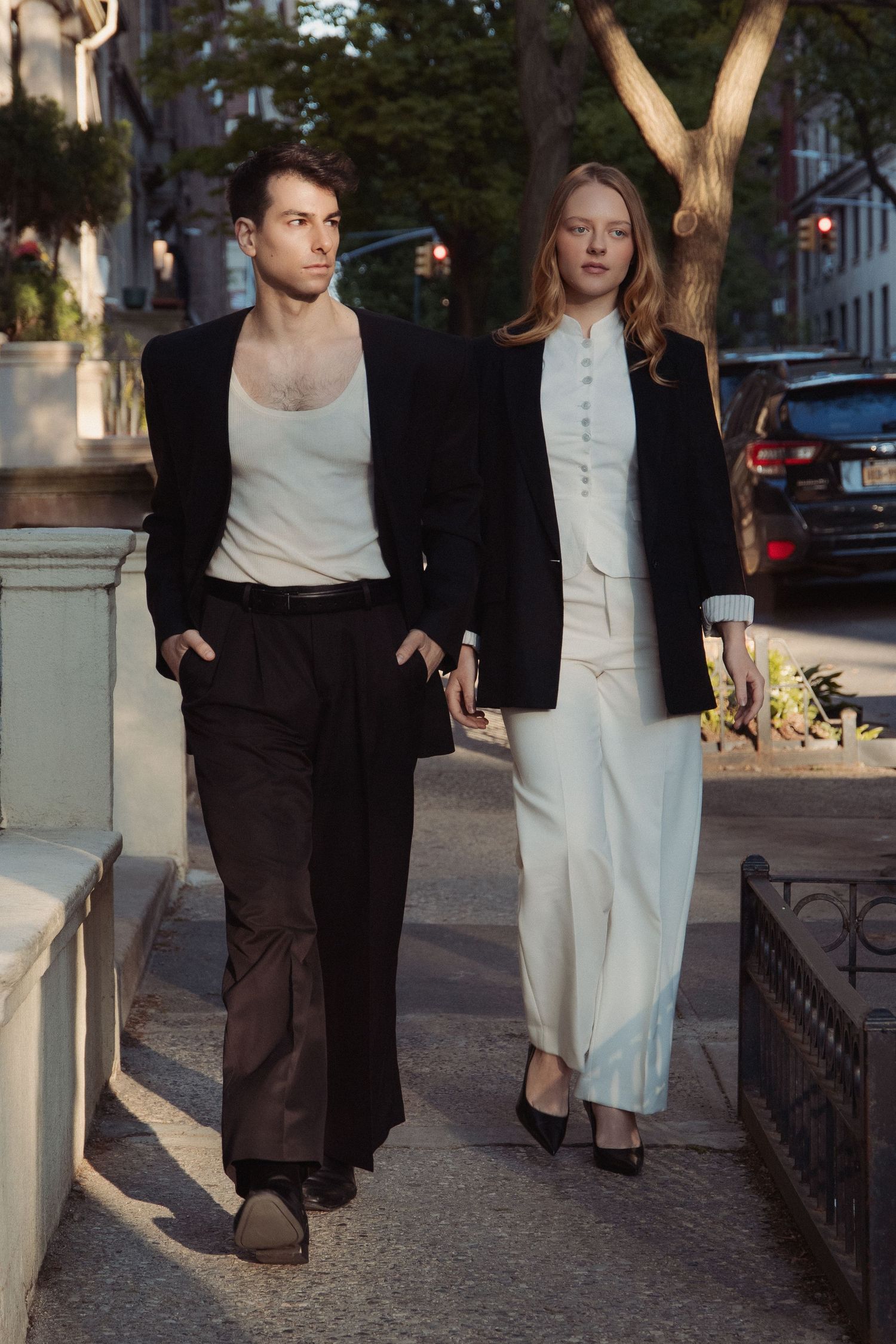 Vin and Regina walk side by side along an Upper West Side sidewalk, their contrasting trousers highlighted by warm late-day sunlight.