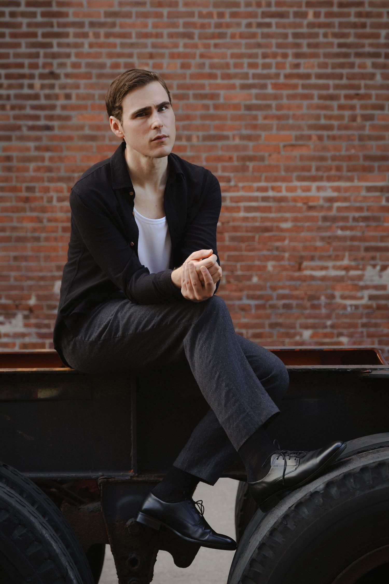 Dylan sits cross-legged on a trailer frame between oversized truck tires, framed closely against a red brick wall.