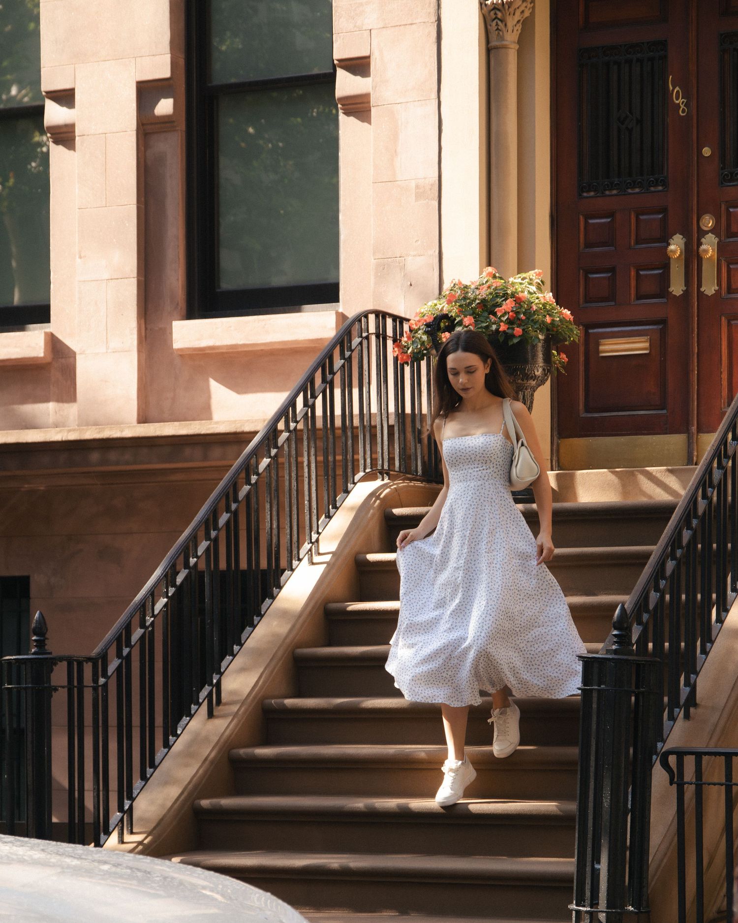 Woman walking down sunlit townhouse steps, holding the sides of her floral dress beneath a flowering planter.