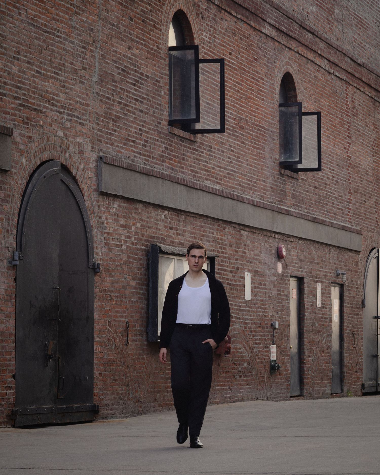 Dylan walks alone across an open industrial courtyard beside a towering red brick warehouse in Red Hook, Brooklyn.