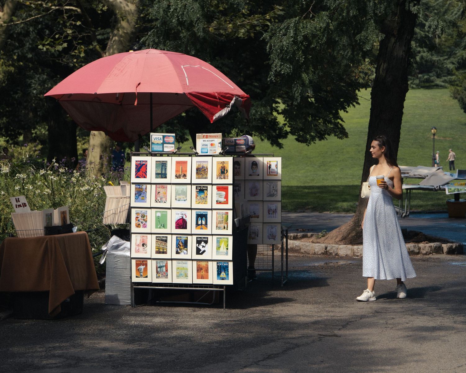 Woman holding coffee beside an outdoor art stall shaded by a red umbrella in Central Park.