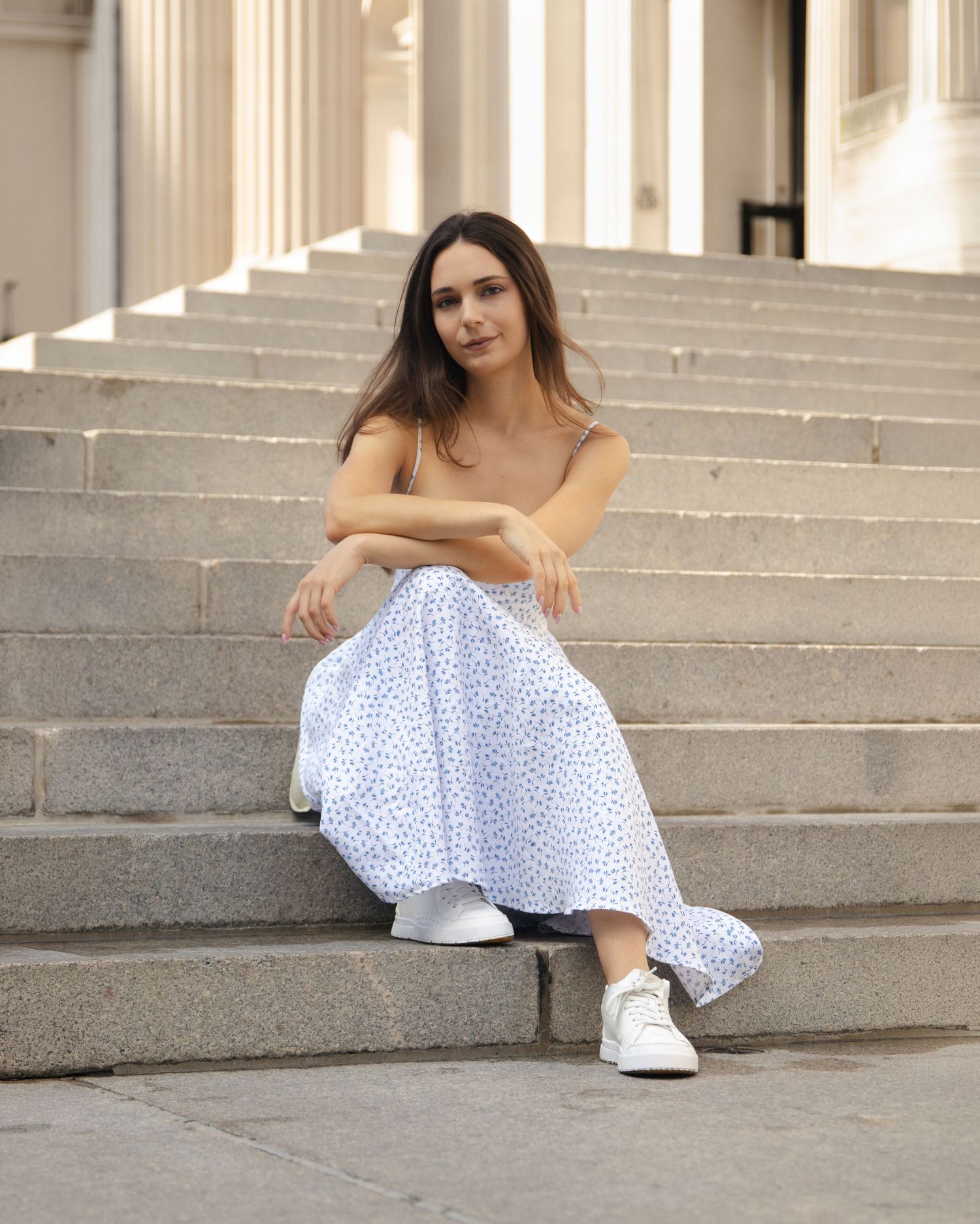 Woman in a floral dress and white sneakers sitting on broad stone steps, with tall columns behind her.