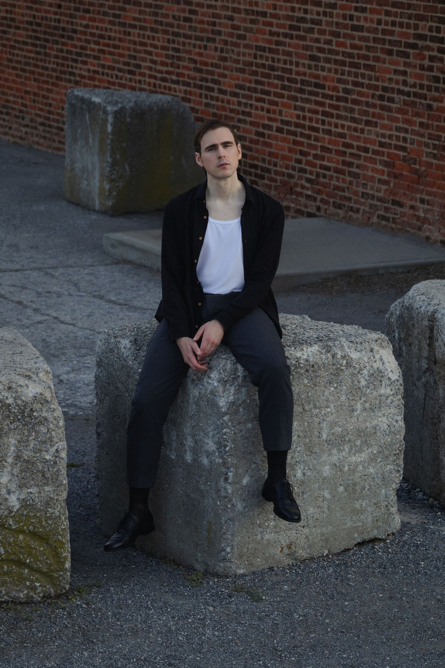 Dylan sits on a large concrete barrier among weathered industrial blocks, with a red brick wall filling the background.
