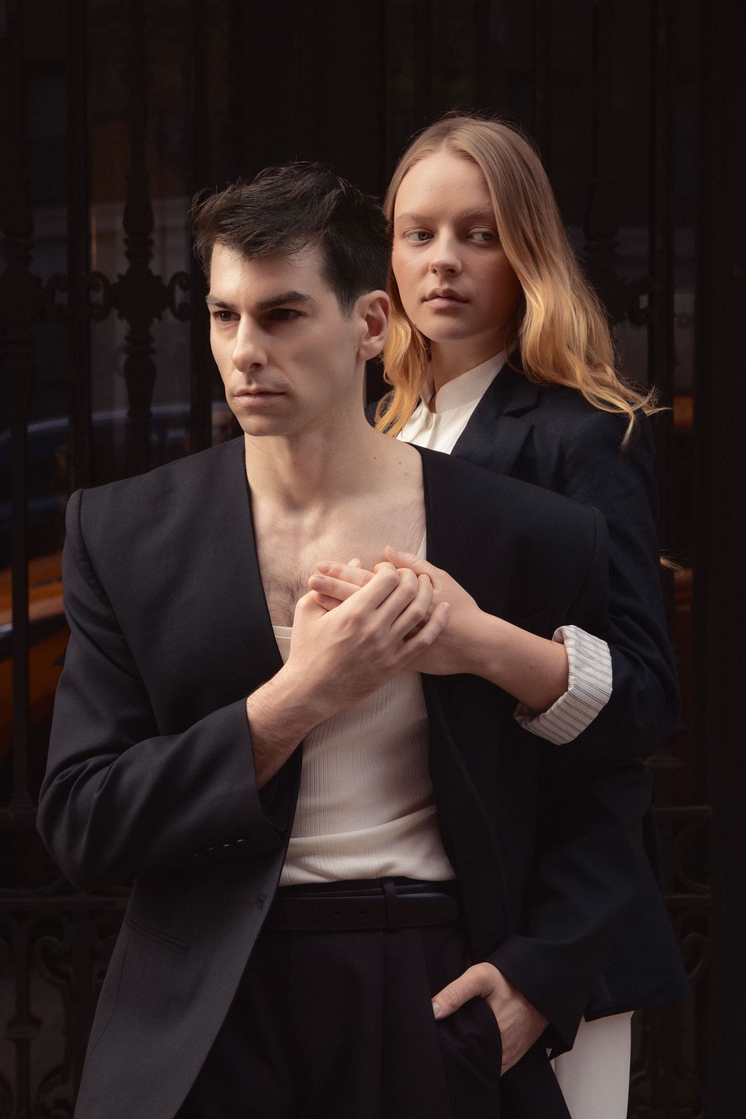 Regina stands behind Vin as they clasp hands in a close editorial portrait against a dark wrought-iron gate.