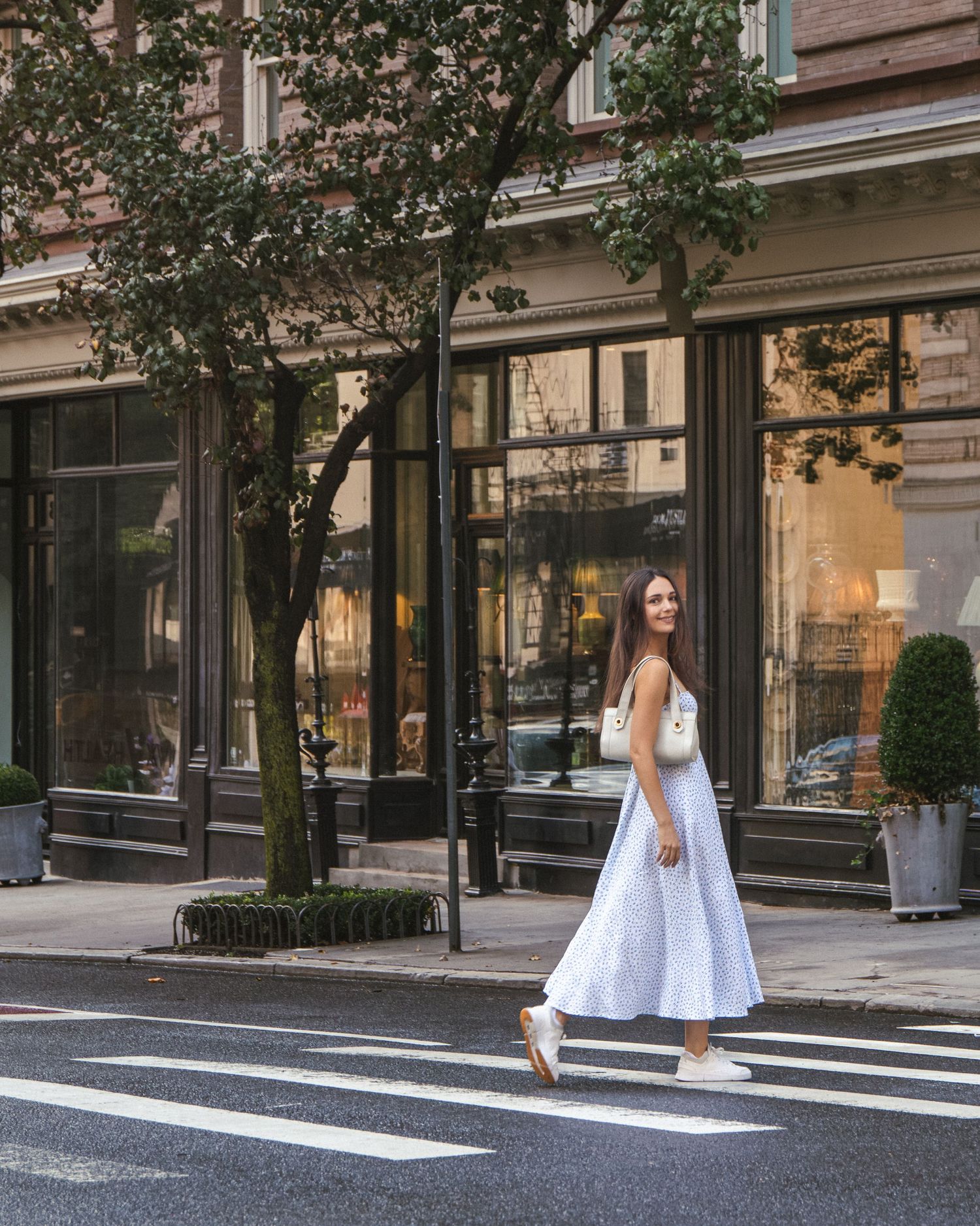 Anastasia in a white floral dress crossing a tree-lined street on Manhattan’s Upper East Side.