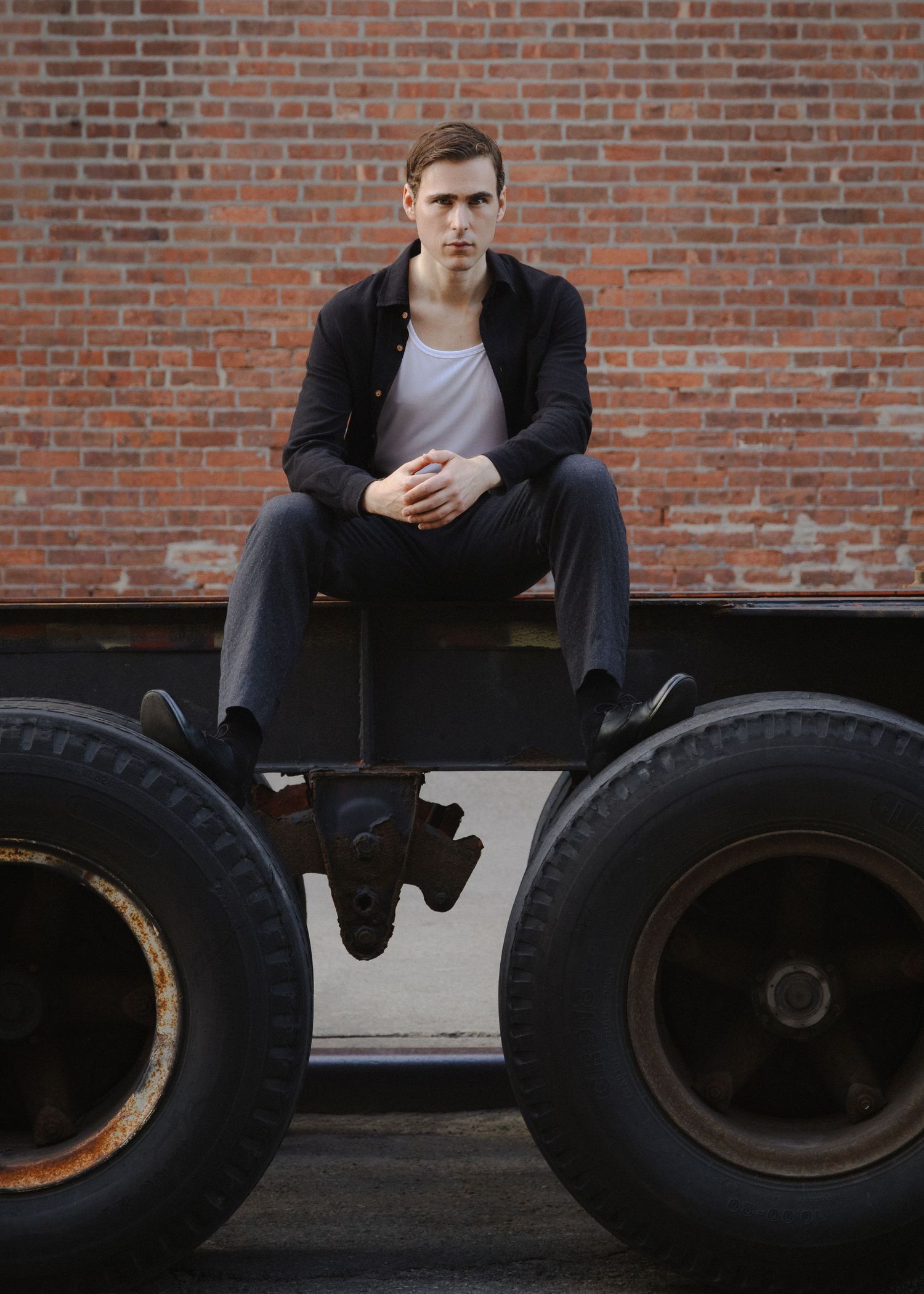 Dylan sits on a metal trailer frame between two oversized truck tires, centered against a red brick wall in Red Hook, Brooklyn.