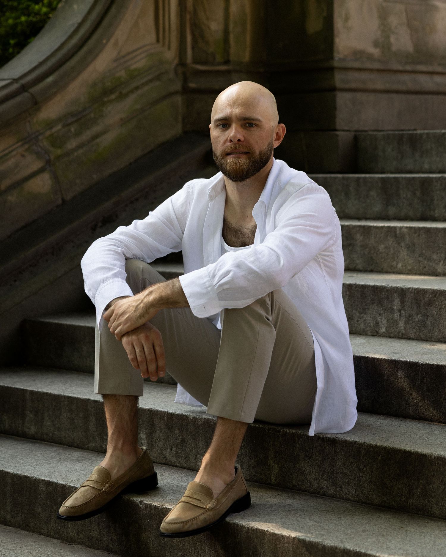 New York City portrait photographer Sergei Kurkin sits on stone steps wearing a white shirt and neutral trousers.