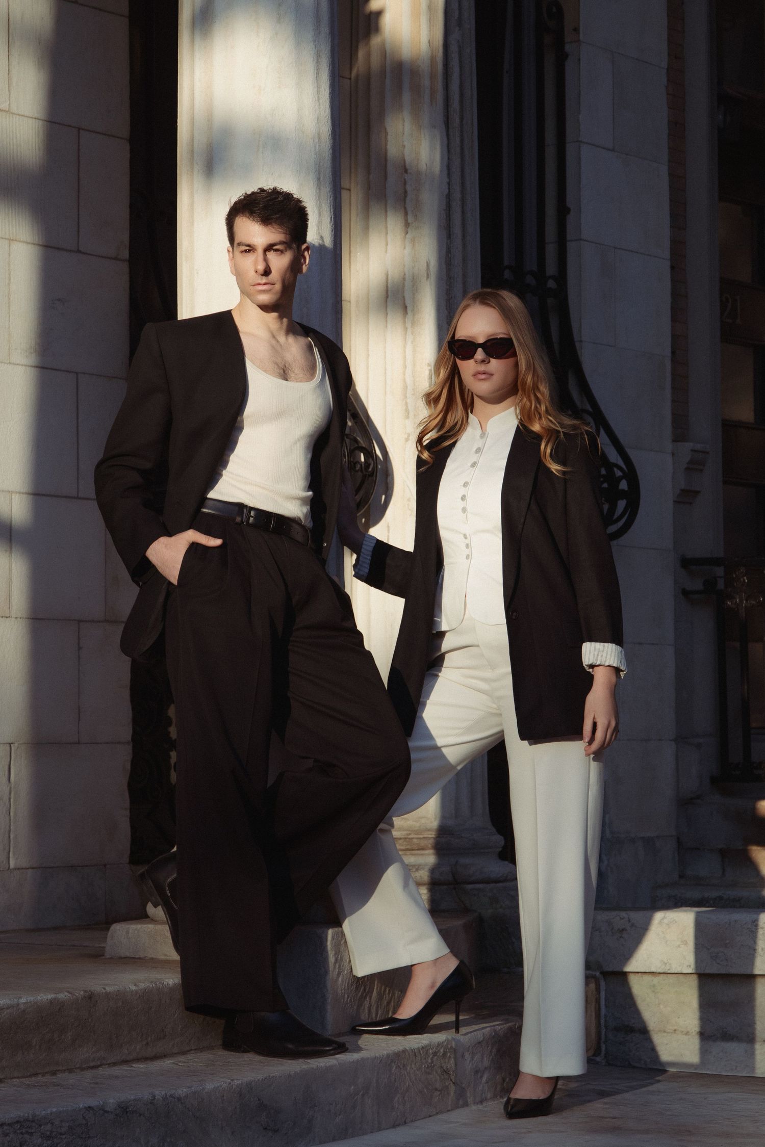 Vin and Regina pose on sunlit stone steps in contrasting black-and-white outfits, with Regina wearing dark sunglasses.