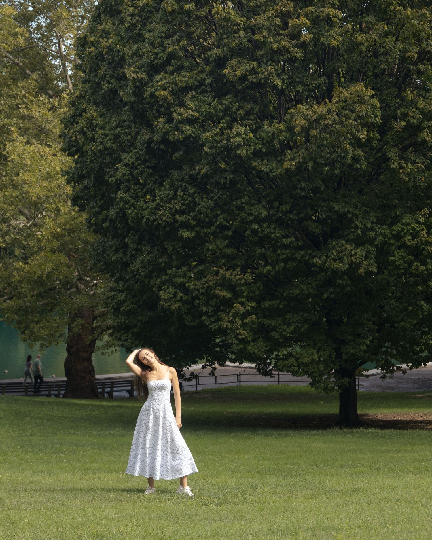 Anastasia standing on a sunlit lawn in Central Park with one hand in her hair, framed by a large leafy tree.