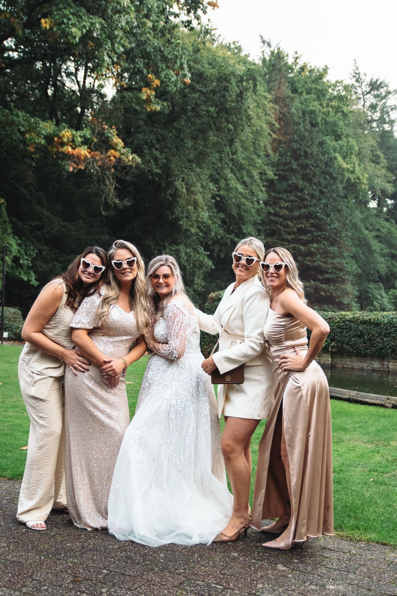 A bride in a white beaded gown poses with four bridesmaids in champagne dresses wearing white sunglasses outdoors.