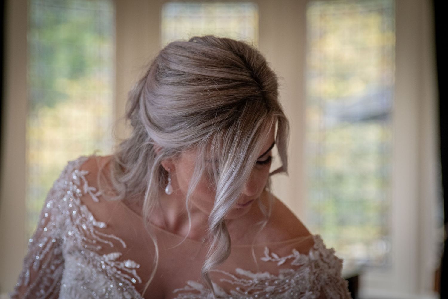 A bride with an elegant updo hairstyle wears an intricate lace wedding gown near a softly lit window.