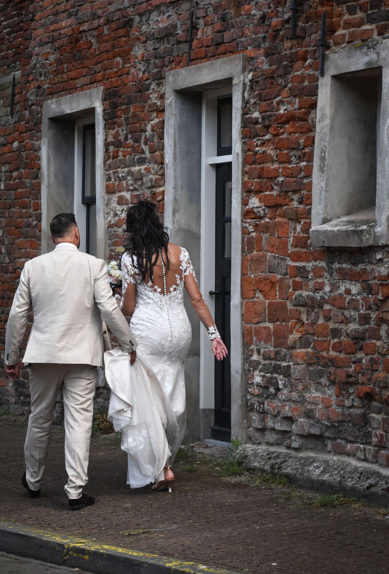 A bride in a white lace gown and groom in a beige suit walk arm in arm along a rustic brick building.