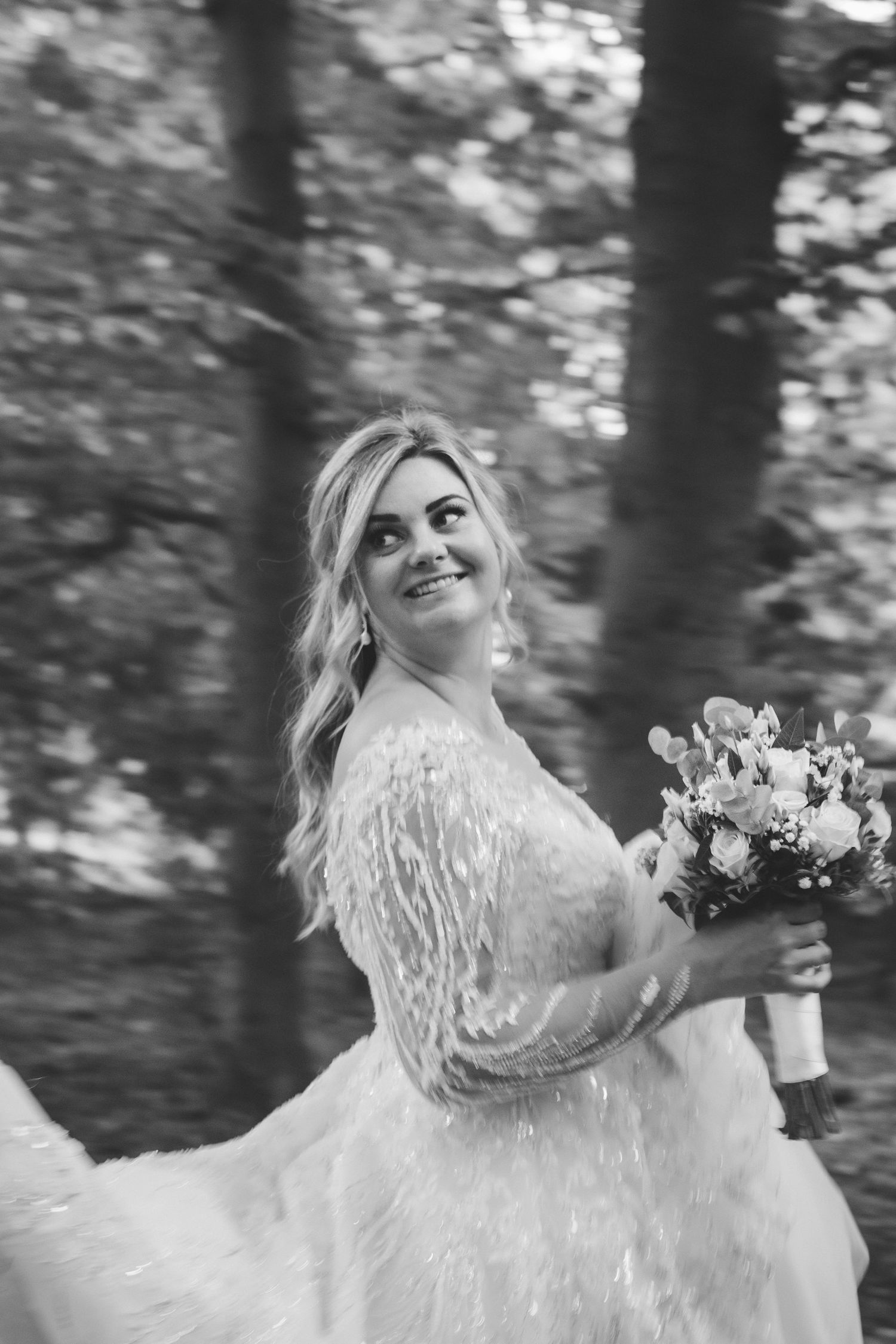 A smiling blonde bride in a feathered white gown holds a floral bouquet in a black and white forest photo.