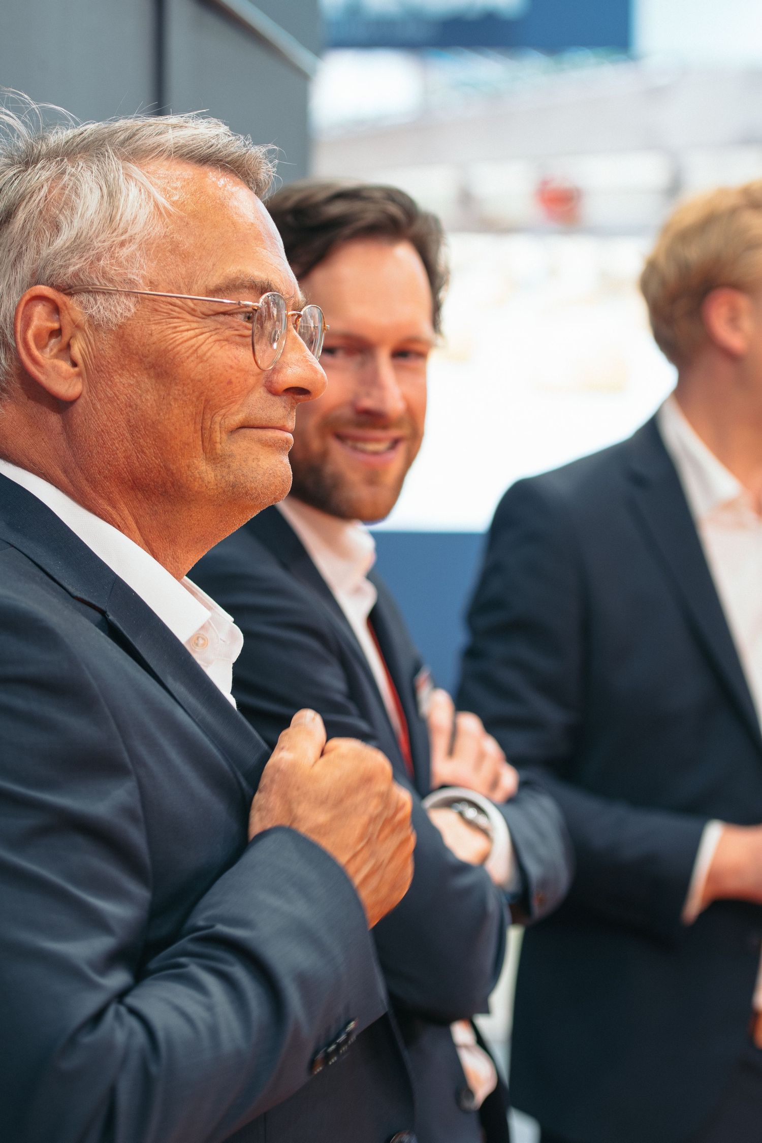 A senior businessman with glasses stands arms crossed with two younger colleagues smiling at a corporate event.