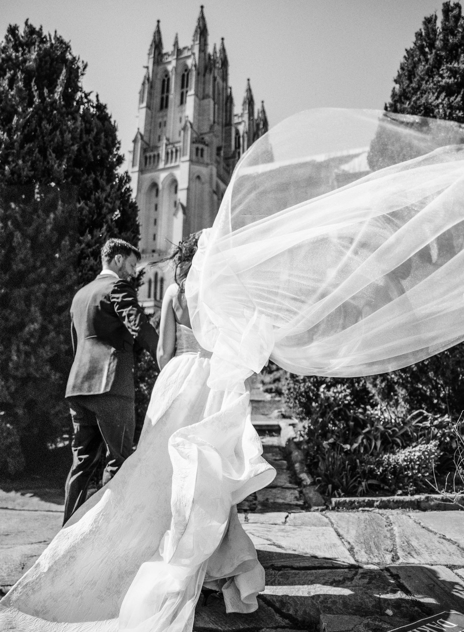 A flowing white wedding veil billows dramatically in the wind against a cathedral backdrop in black and white photography.