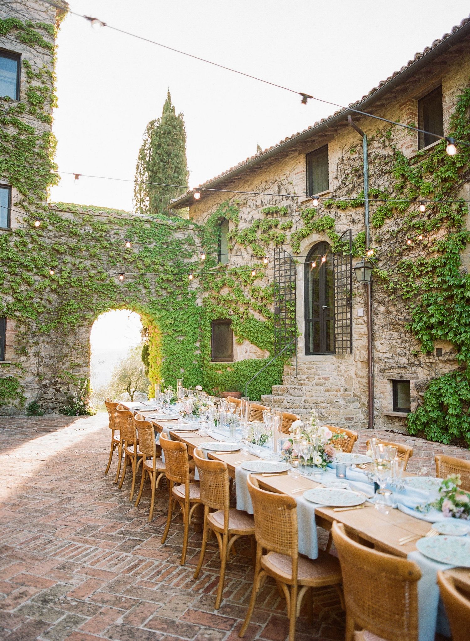 Long rustic dining table set for an elegant outdoor meal on a stone patio surrounded by ivy-covered walls at sunset.