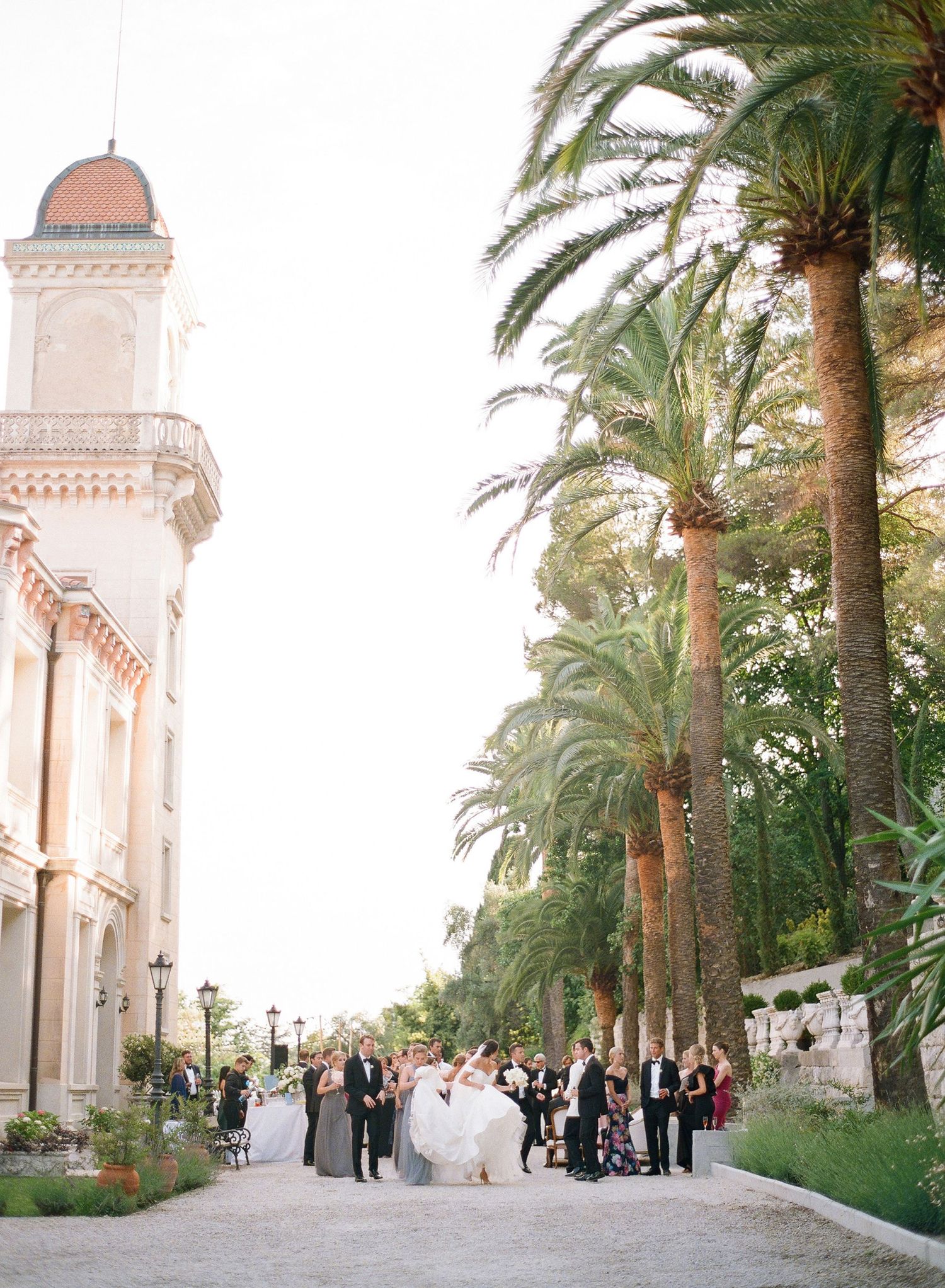 Palm trees line a path leading to an elegant Mediterranean-style church with a pink dome and gathering guests.
