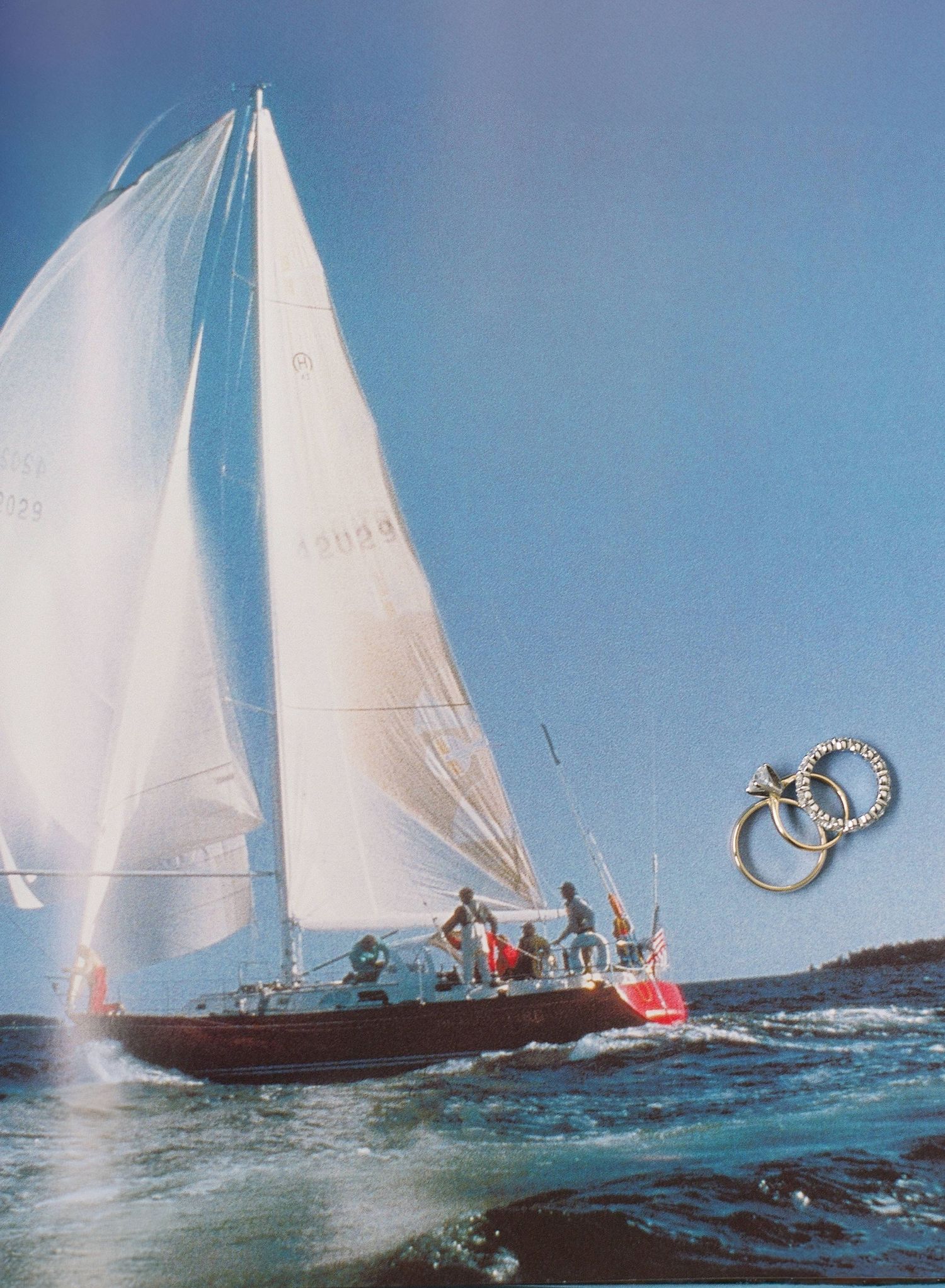 A sailboat with white sails glides across blue waters on a sunny day with crew members visible on deck.