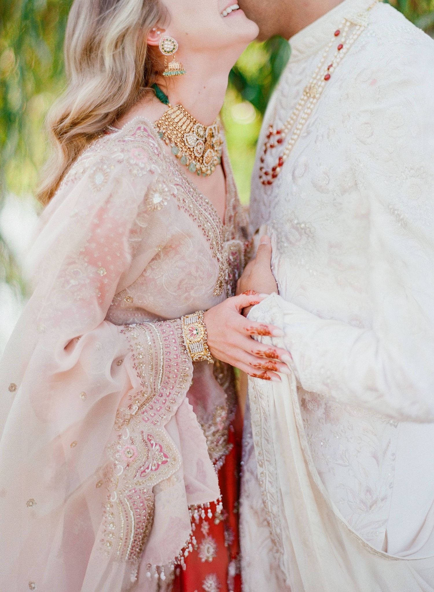 Couple in traditional South Asian wedding attire embrace wearing ornate pink and cream outfits with gold jewelry.
