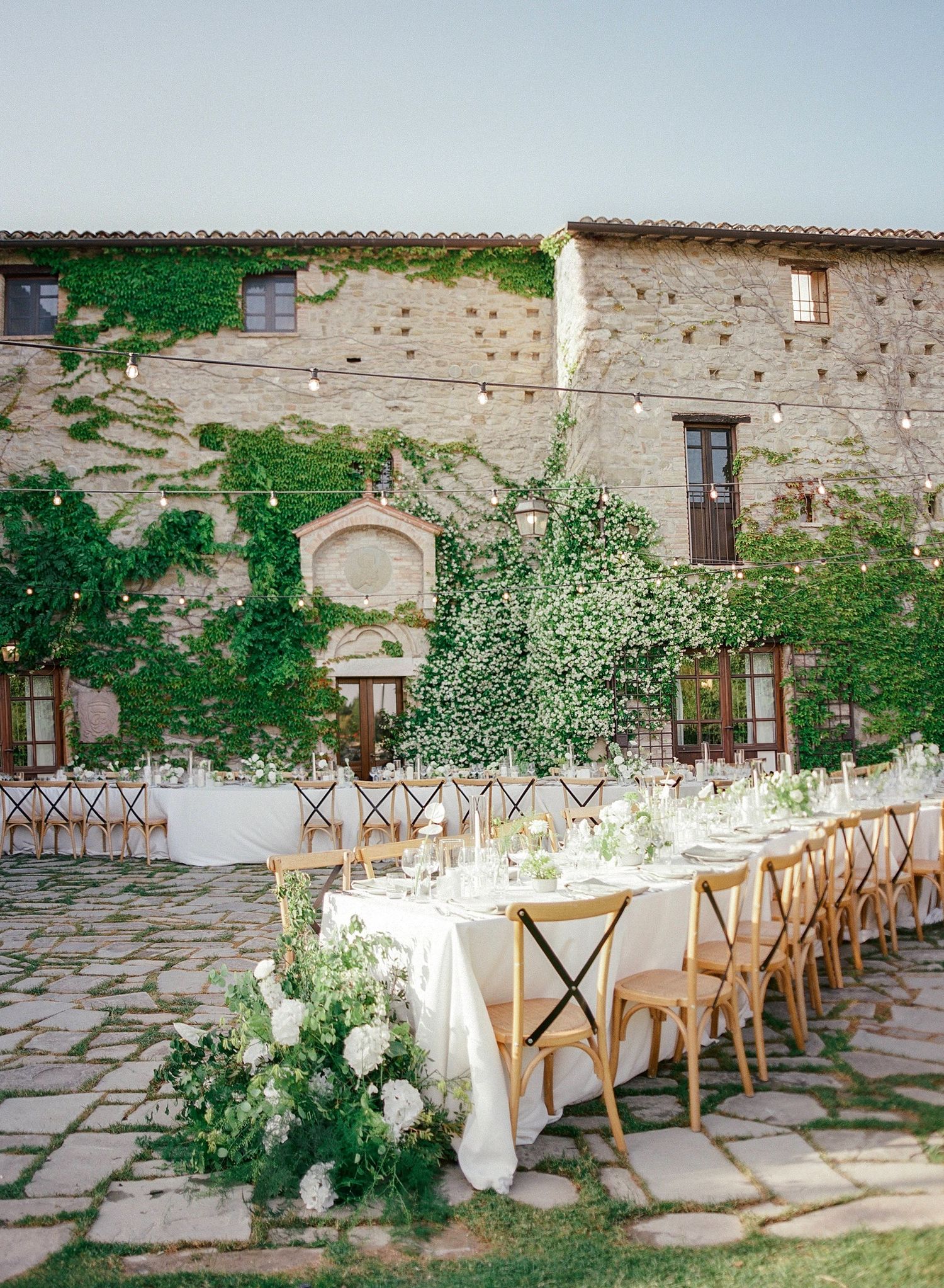 Elegant outdoor wedding reception table setup against rustic stone villa with climbing vines and white floral arrangements.