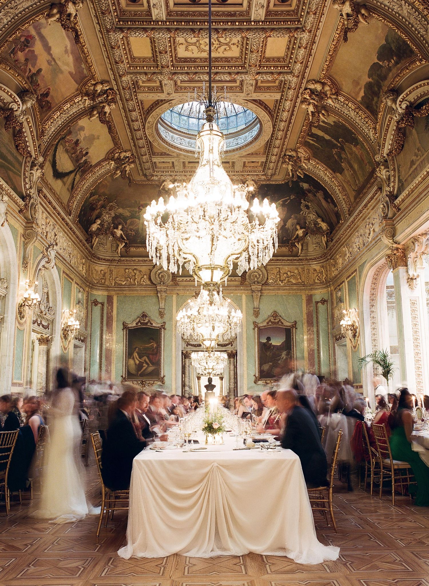 Elegant wedding reception in an ornate ballroom with crystal chandeliers and gilded details showing multiple table settings.