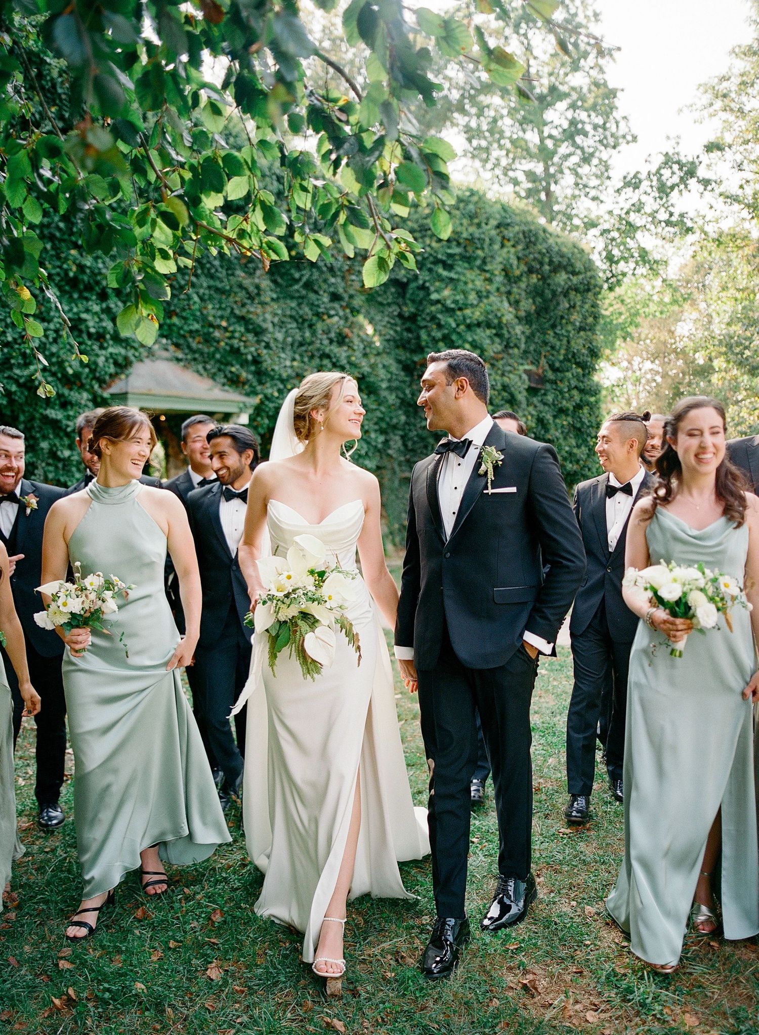 Wedding party in formal attire walks together outdoors in a candid moment along a tree-lined path.