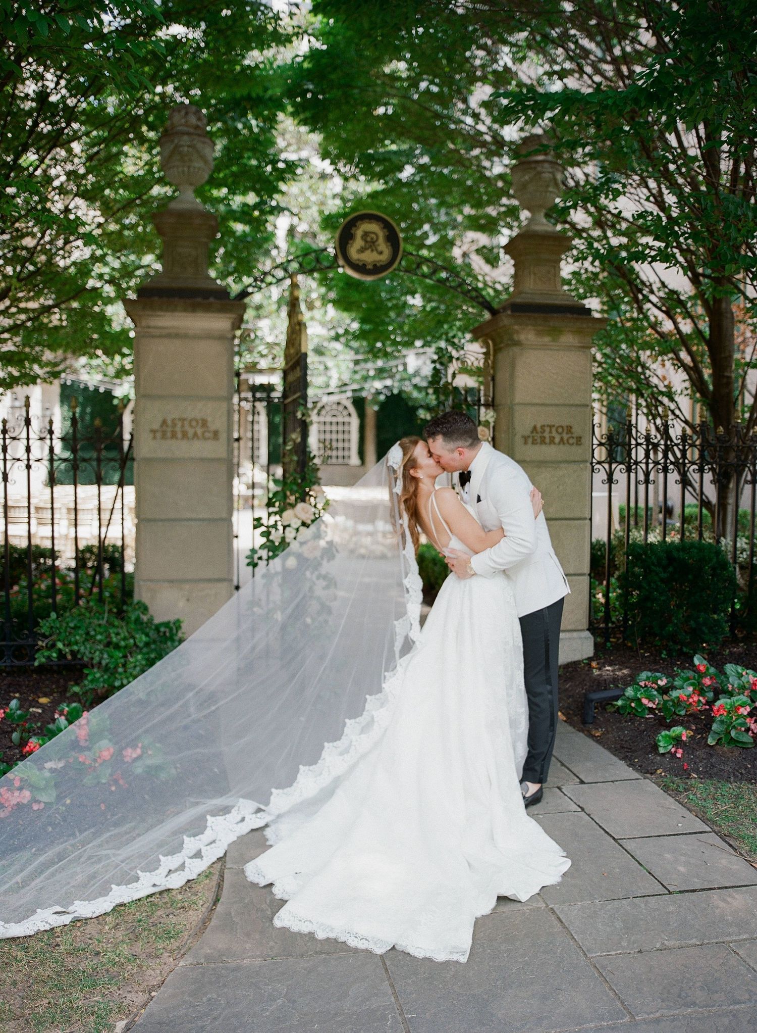 A romantic wedding couple shares an intimate kiss in a garden courtyard with a flowing white veil and classic architecture.
