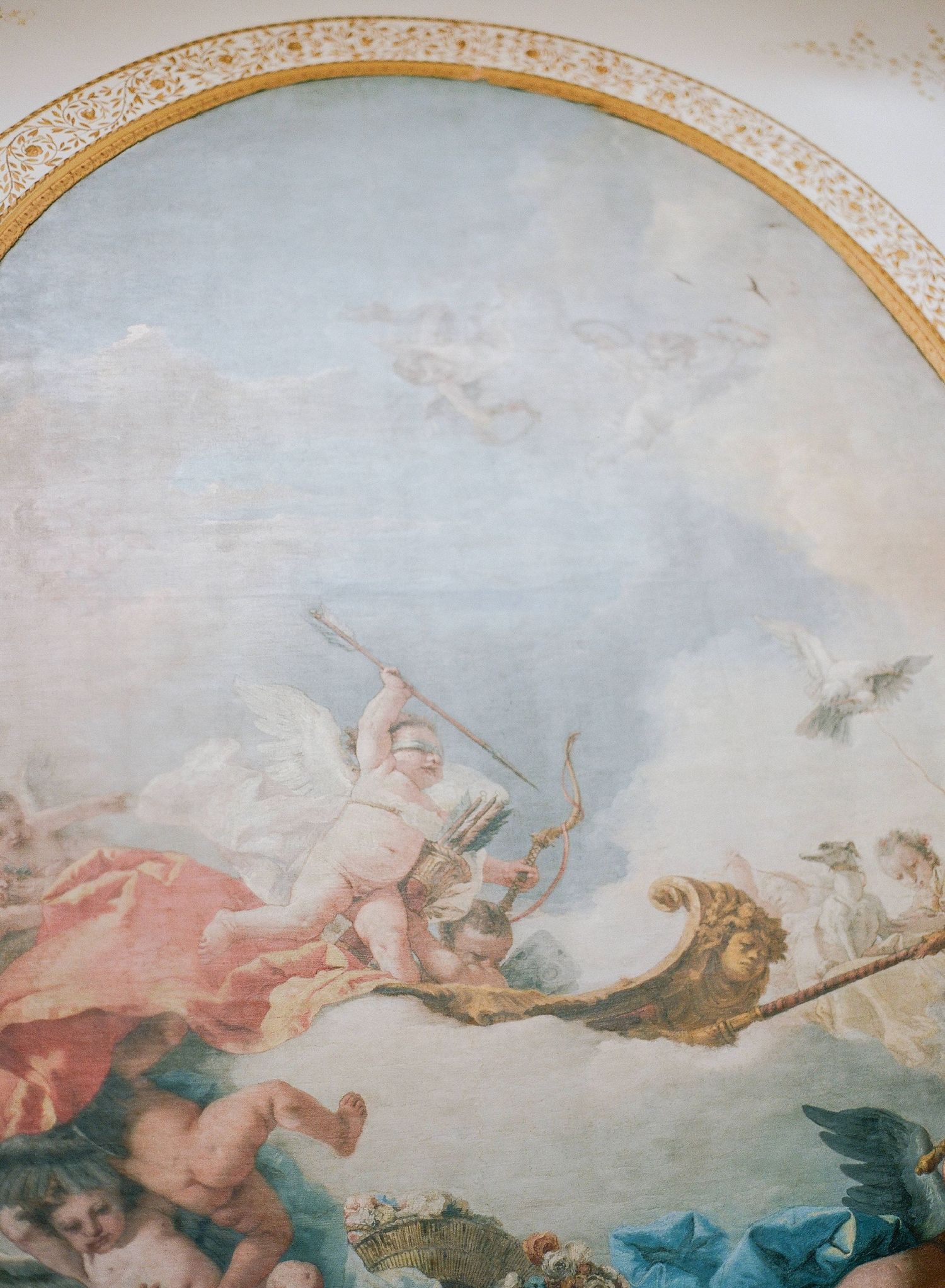Baroque ceiling fresco depicting cherubs and mythological figures soaring through cloudy skies with ornate golden frame details.