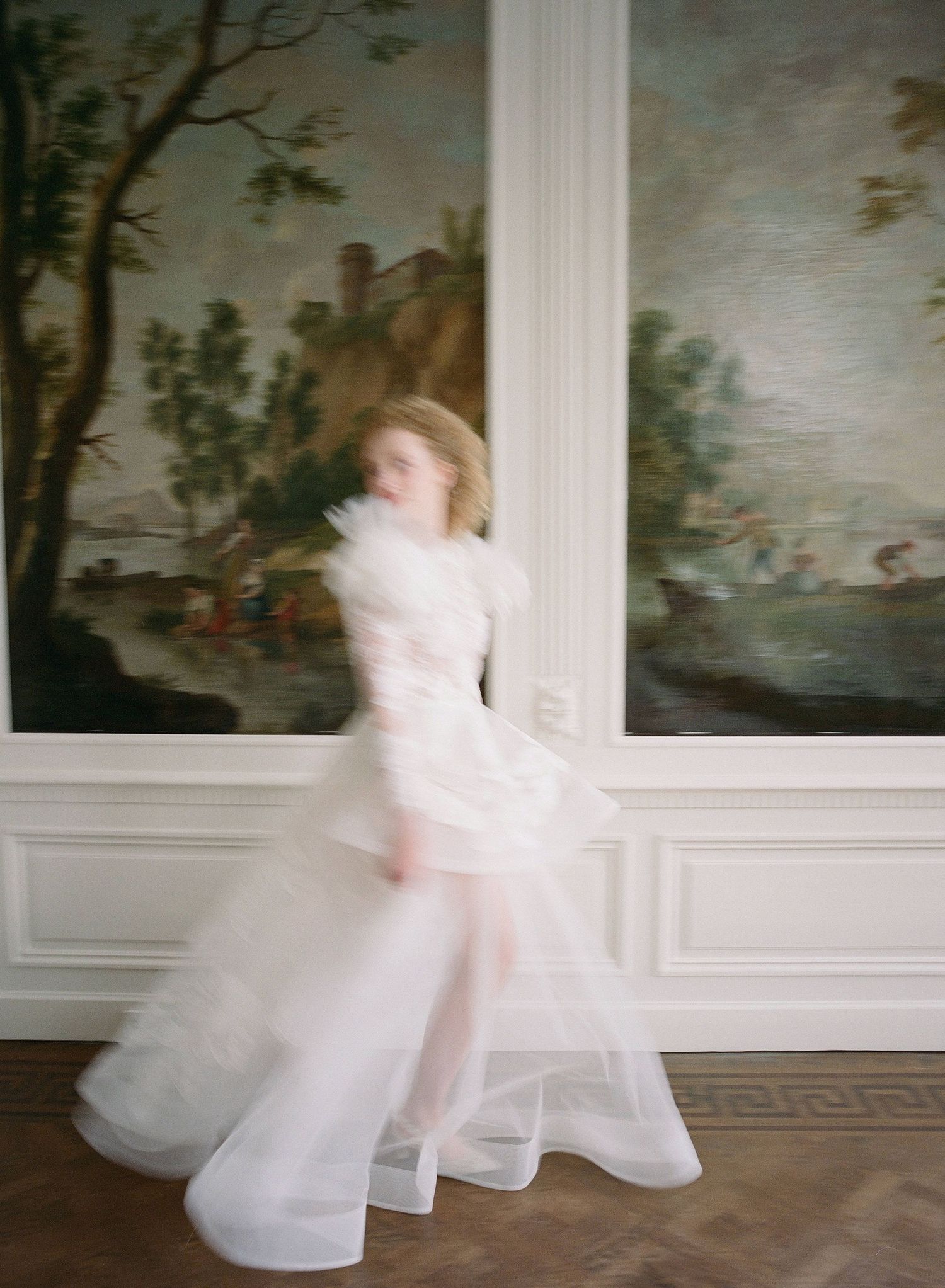 A blurred figure in a flowing white gown twirls and dances in an elegant room with painted wall murals.