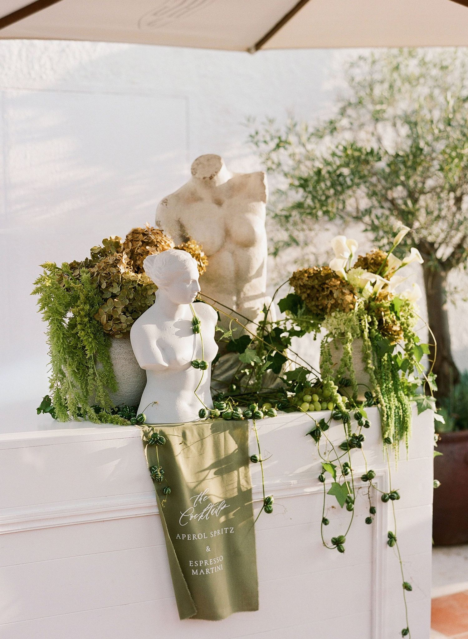 White cherub statue and lush greenery arrangements decorate an elegant outdoor event space under a canopy.