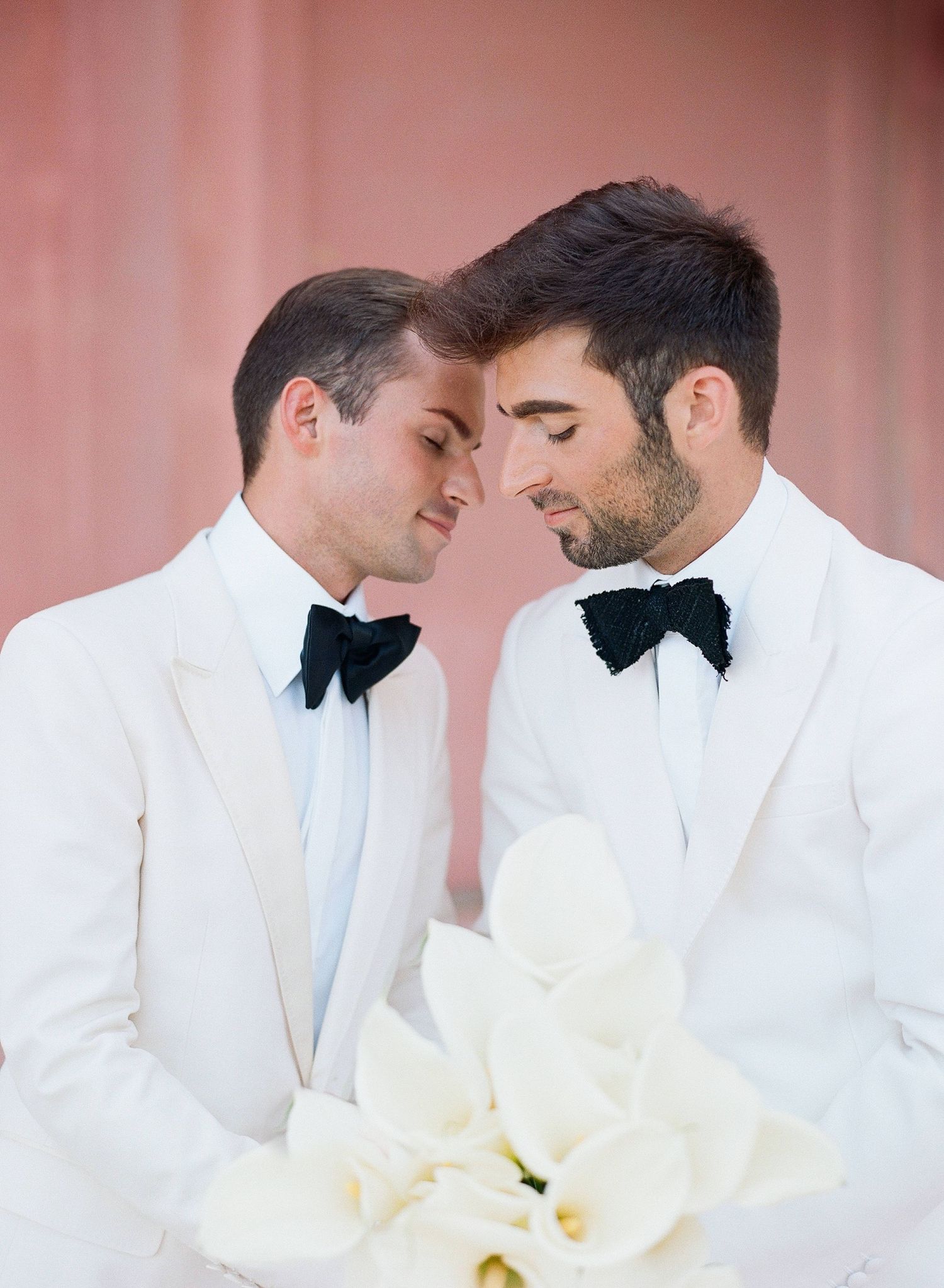 Two people in white suits share an intimate moment against a soft pink background during a wedding celebration.