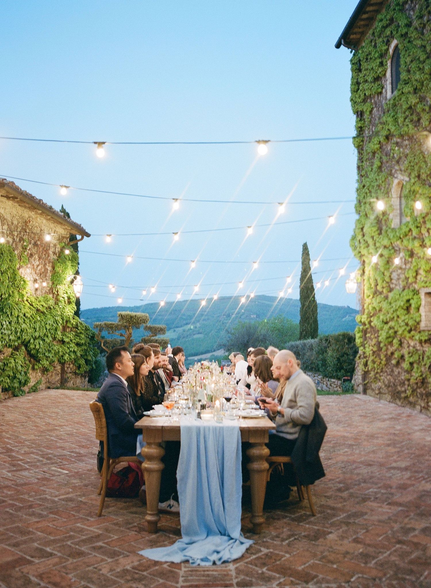 Elegant outdoor wedding reception setup with string lights and blue tablecloths on a rustic stone terrace overlooking mountains.
