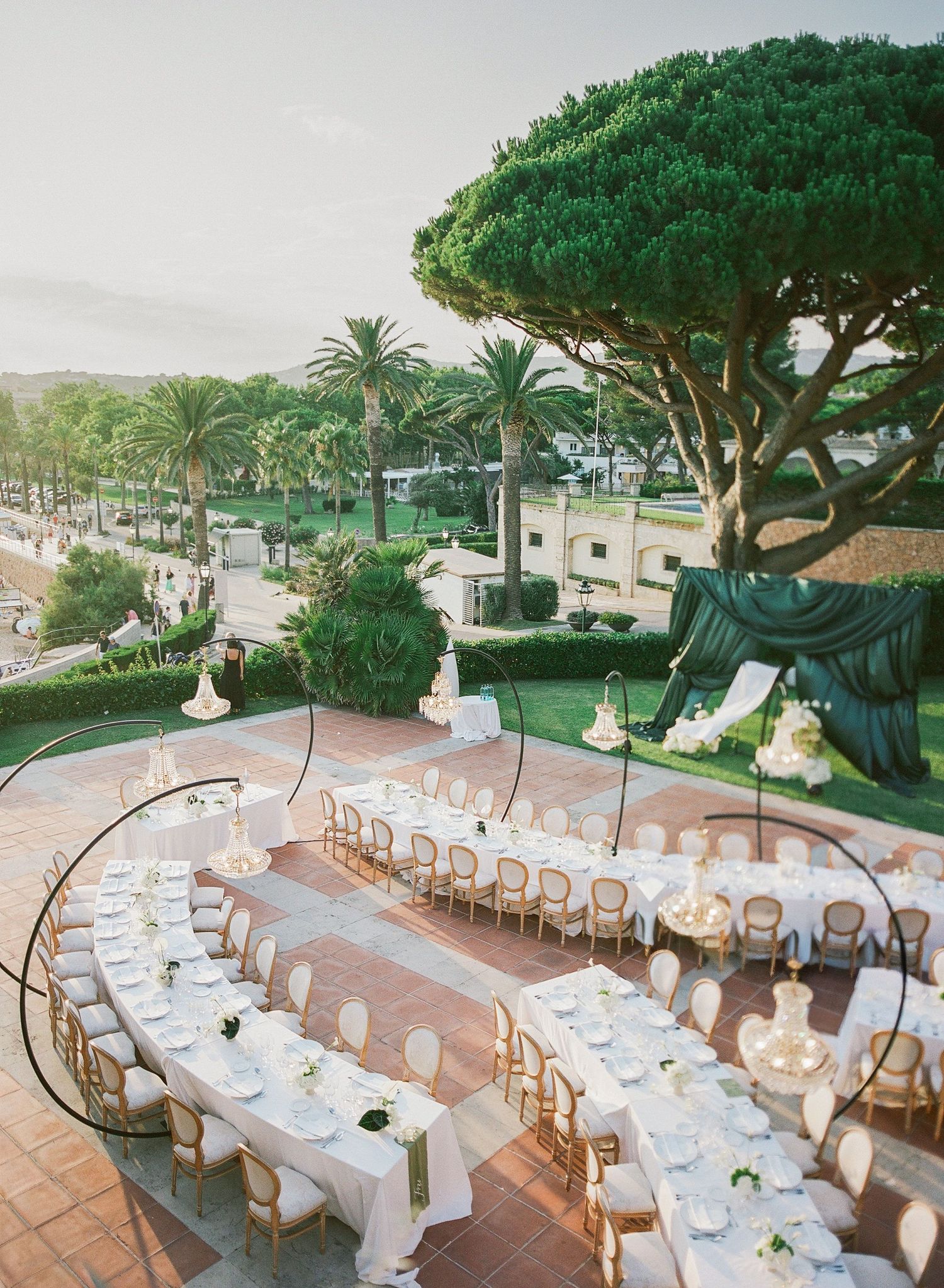 Elegant outdoor wedding reception setup with long white tables and wooden chairs under lush green trees and palm trees.