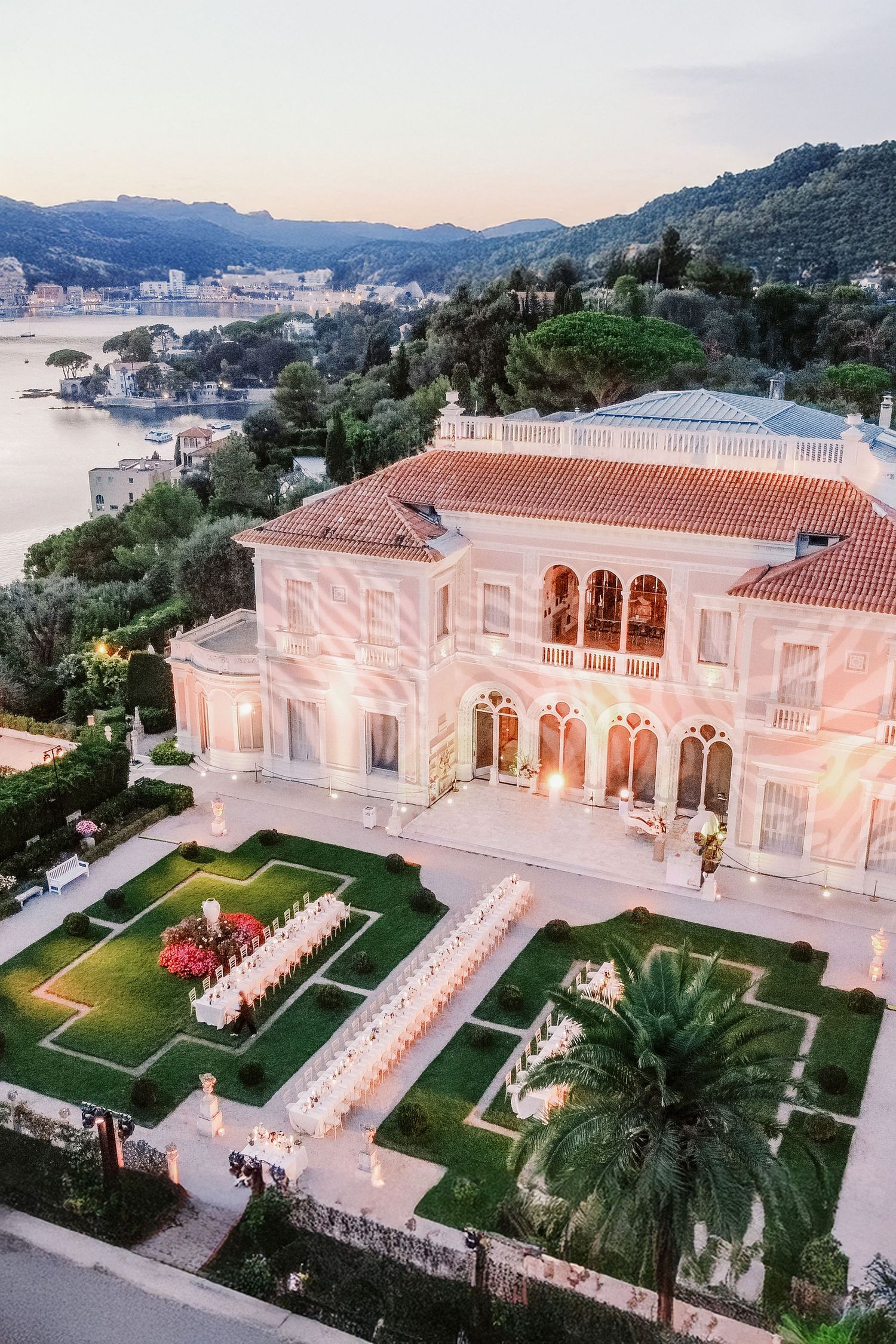 Luxurious white Mediterranean villa with illuminated terraces and palm trees overlooking a coastal bay at sunset.