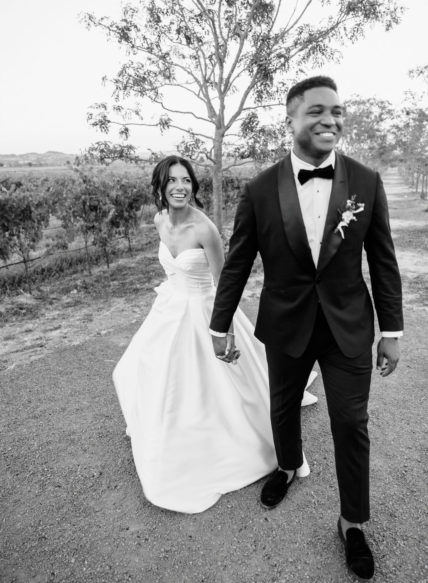 A joyful couple in wedding attire walks and laughs together on a grassy hillside path in black and white.