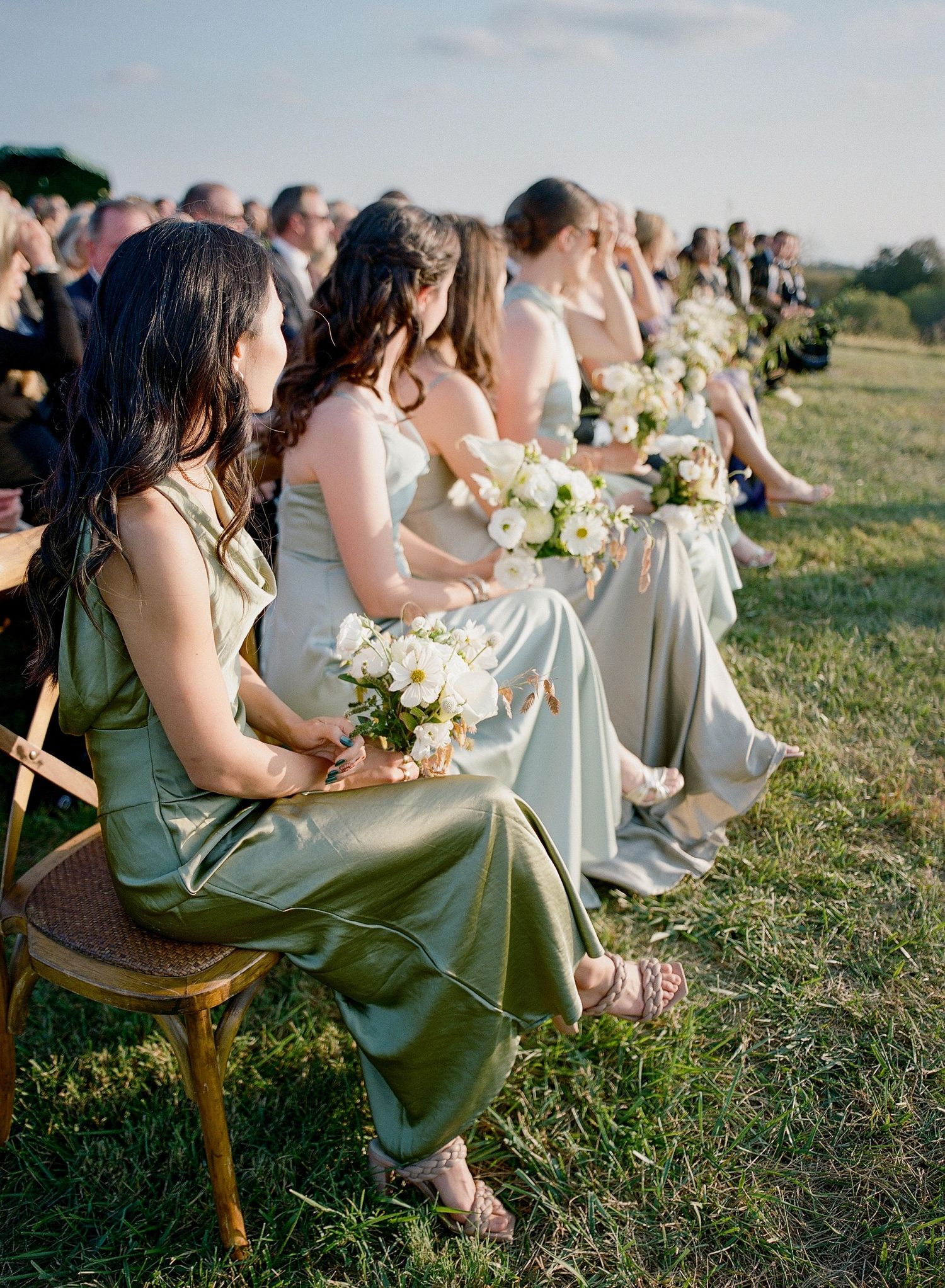 Bridesmaids in sage green dresses sit on vintage wooden chairs in a grassy outdoor setting.