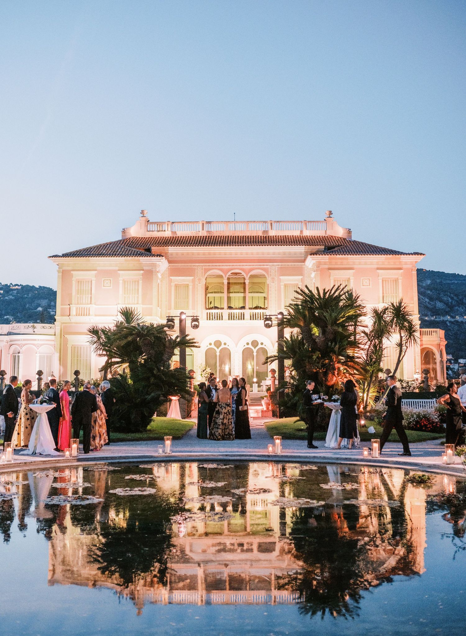 Luxurious pink villa with palm trees reflecting in still water during sunset at a Mediterranean coastal estate.