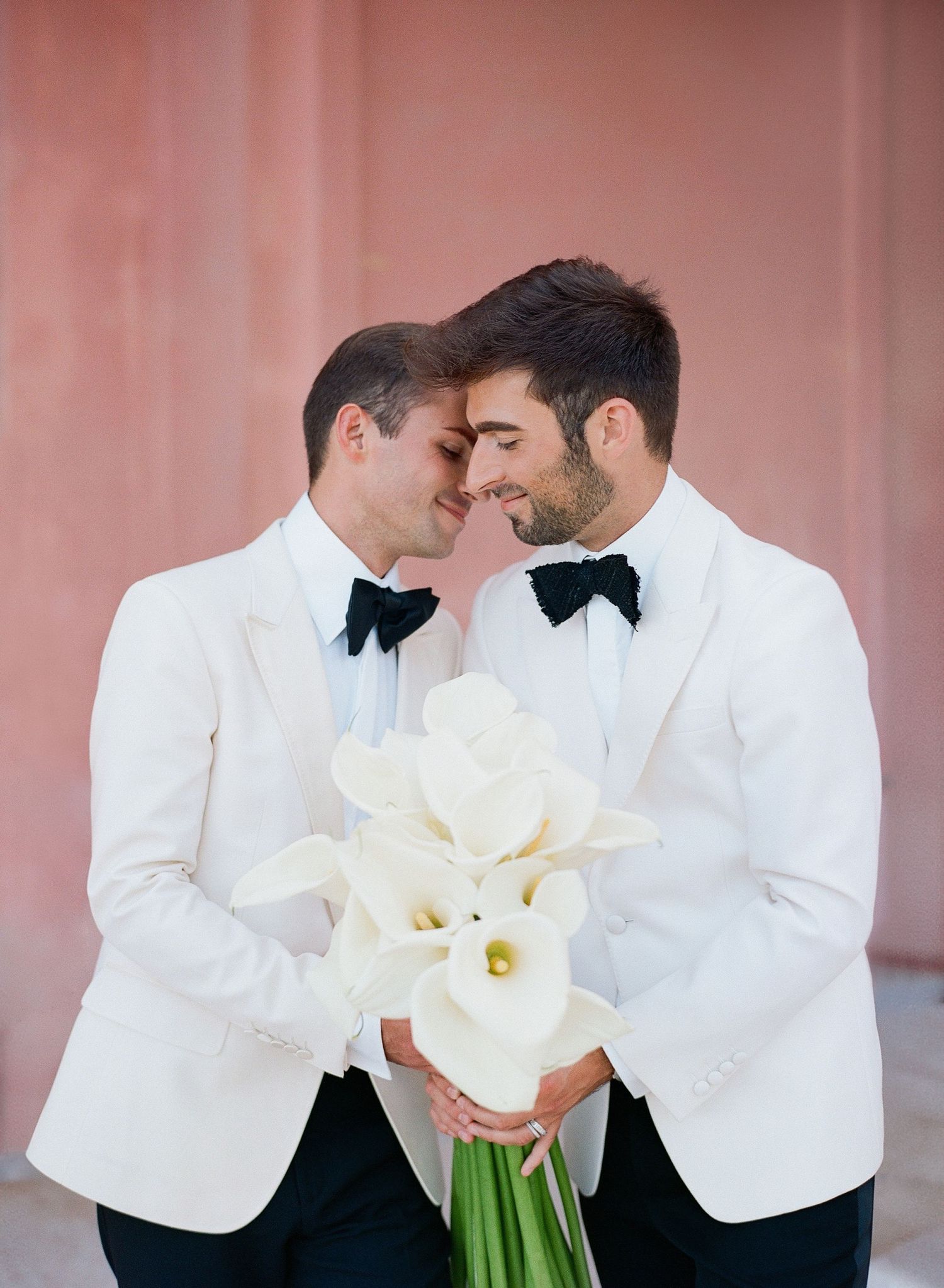 Two people in white tuxedo jackets and black bow ties share an intimate moment while holding white calla lily bouquet.