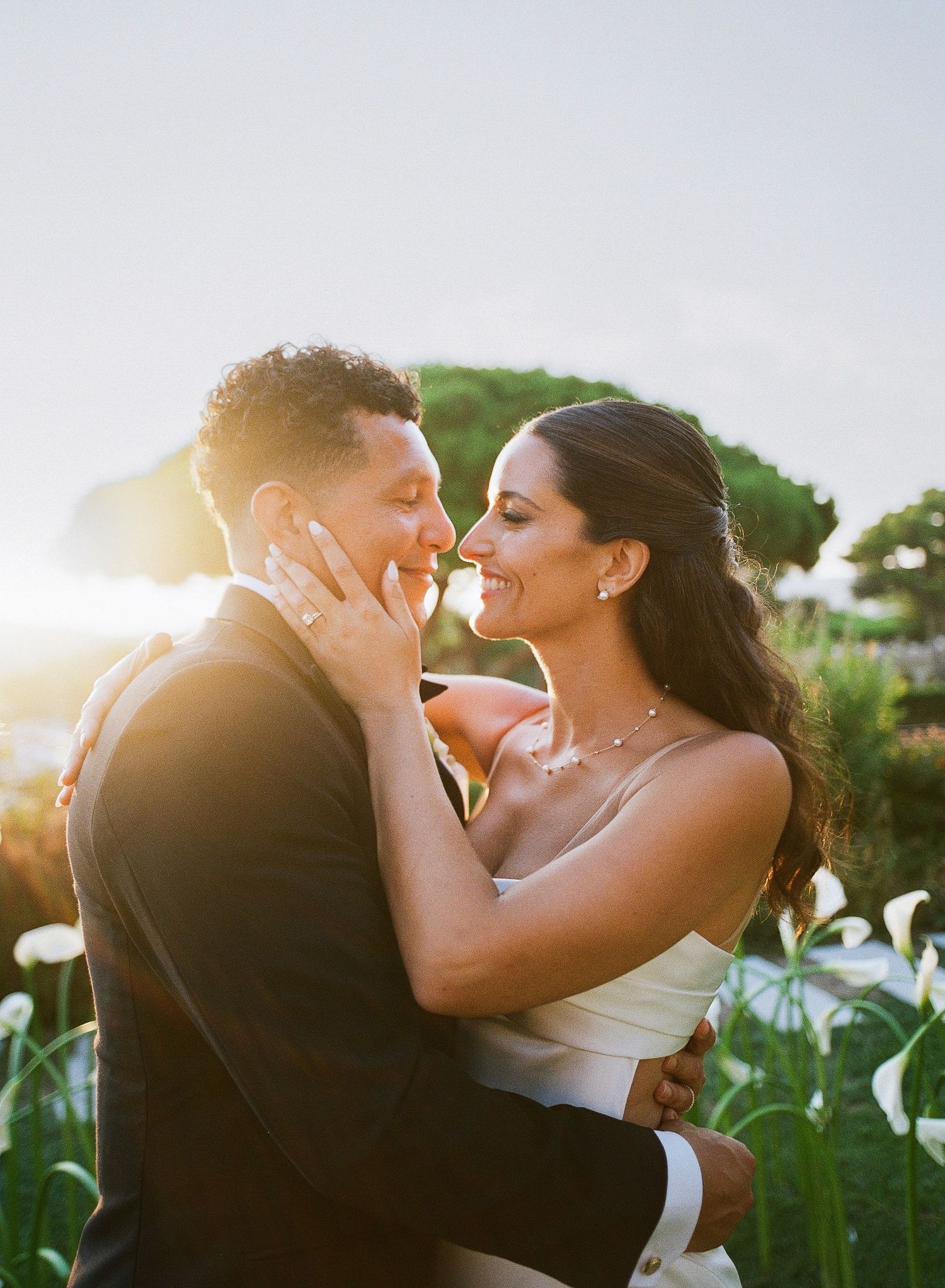 Romantic wedding couple sharing intimate kisses at sunset with lush greenery and golden light in background.