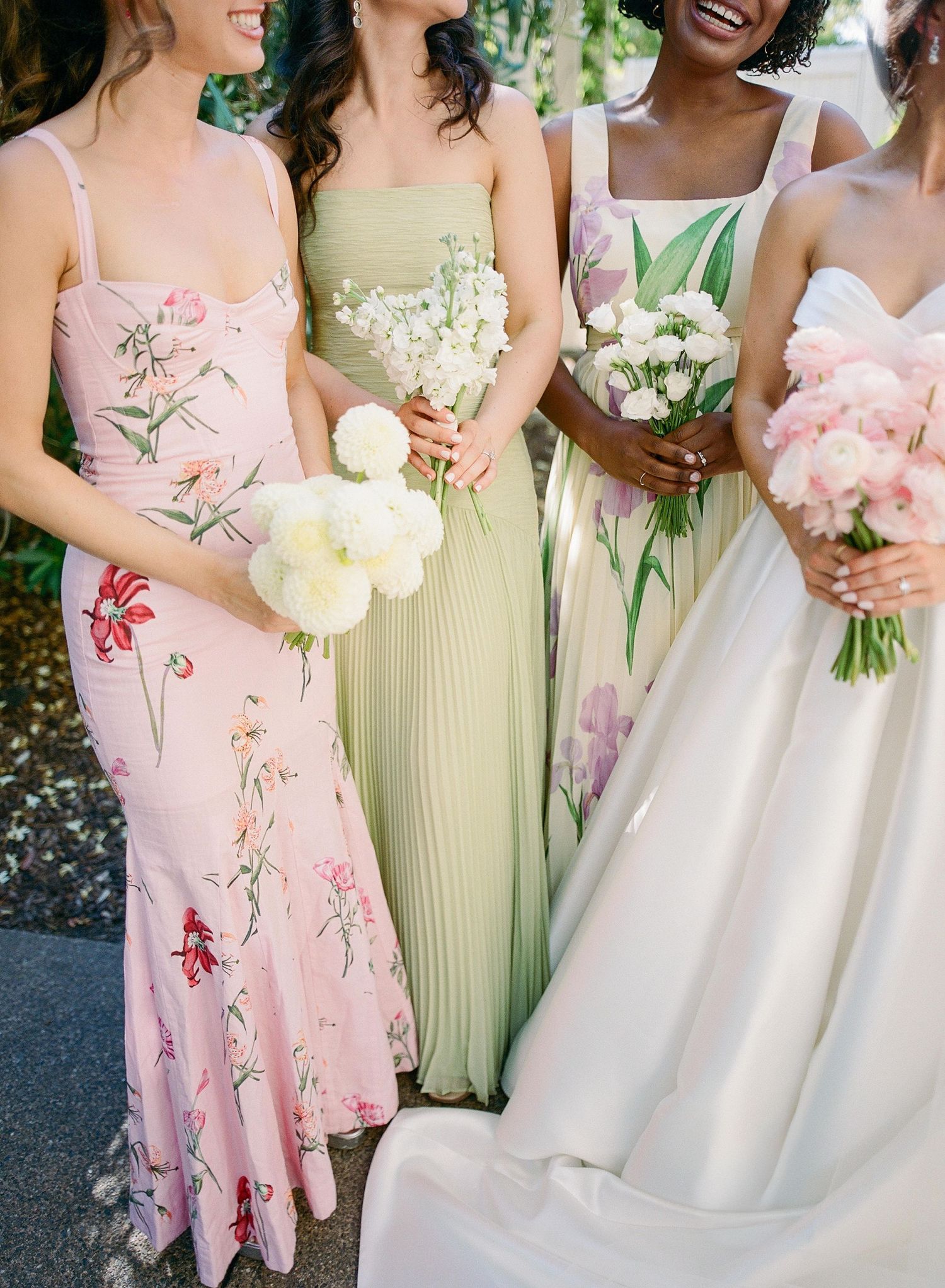 Bridesmaids in soft pink floral and pale yellow dresses hold pastel bouquets at an outdoor wedding celebration.