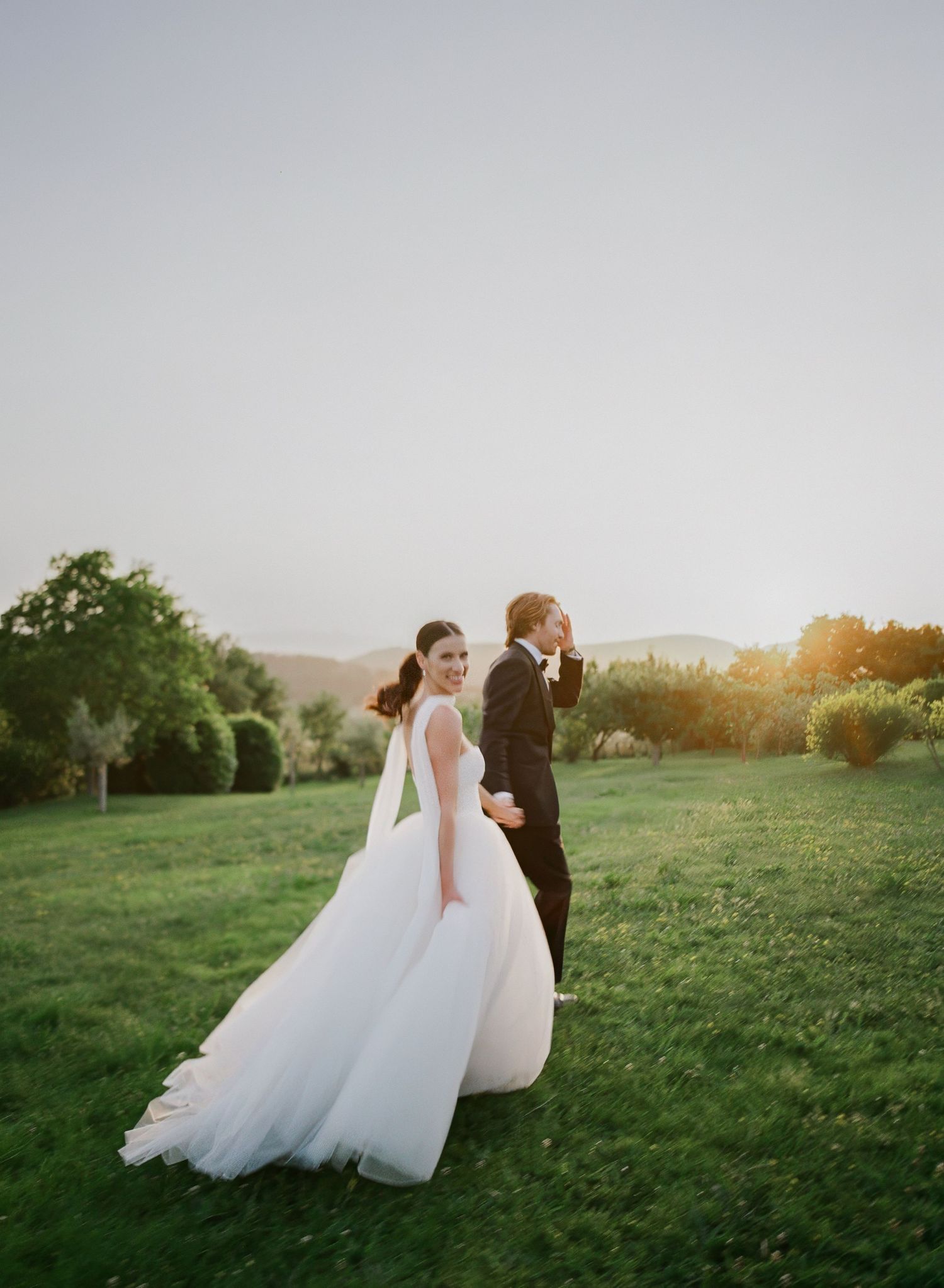 A couple in wedding attire walks hand in hand through a sunlit meadow at golden hour.