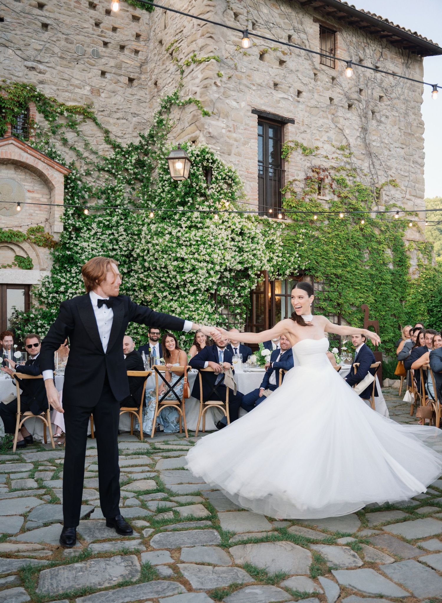 A couple performs a romantic twirling dance sequence in front of stone walls at an outdoor wedding venue.