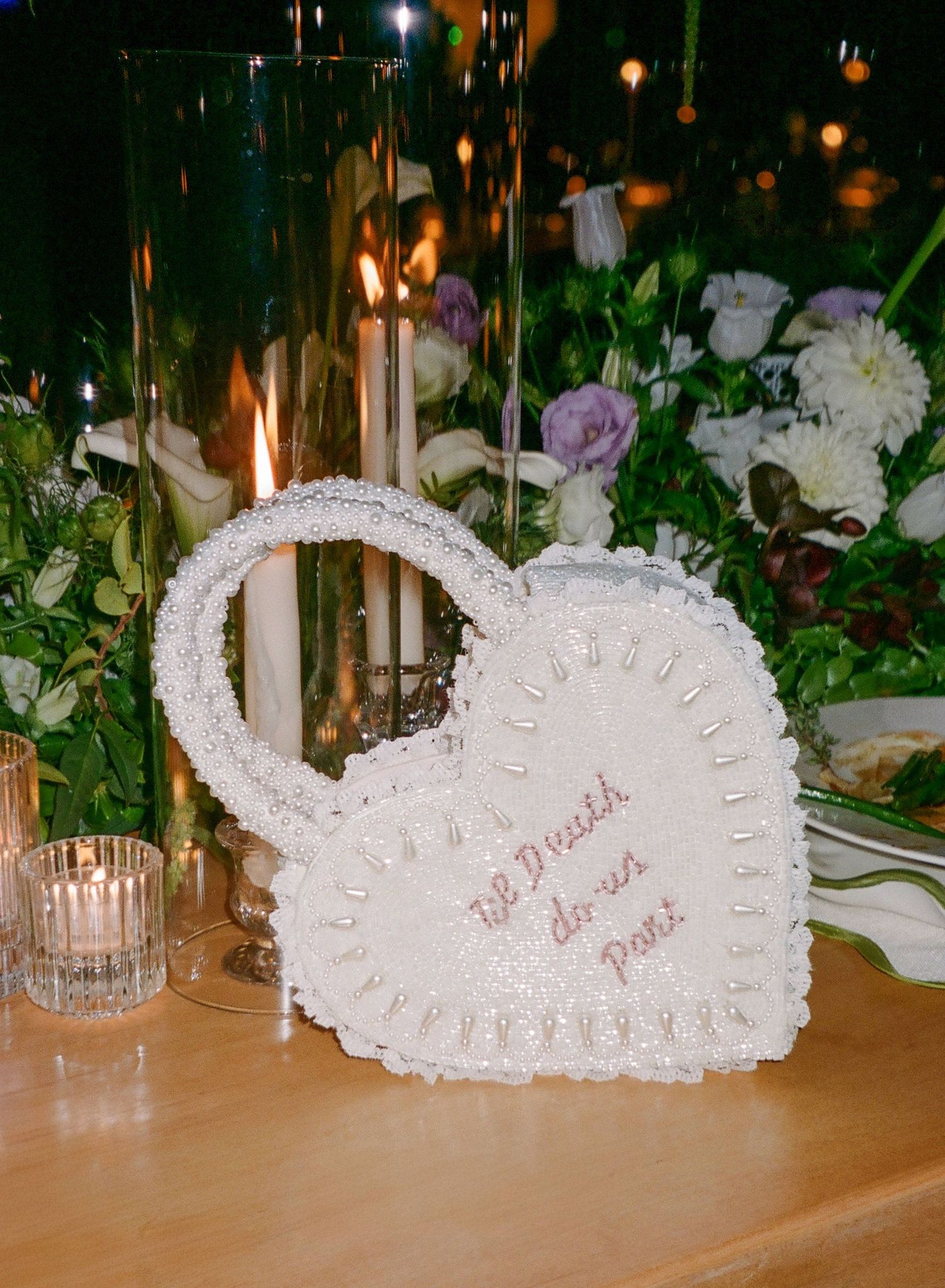 White lace heart-shaped decorative piece with candles and purple and white flowers on a wooden table at night.