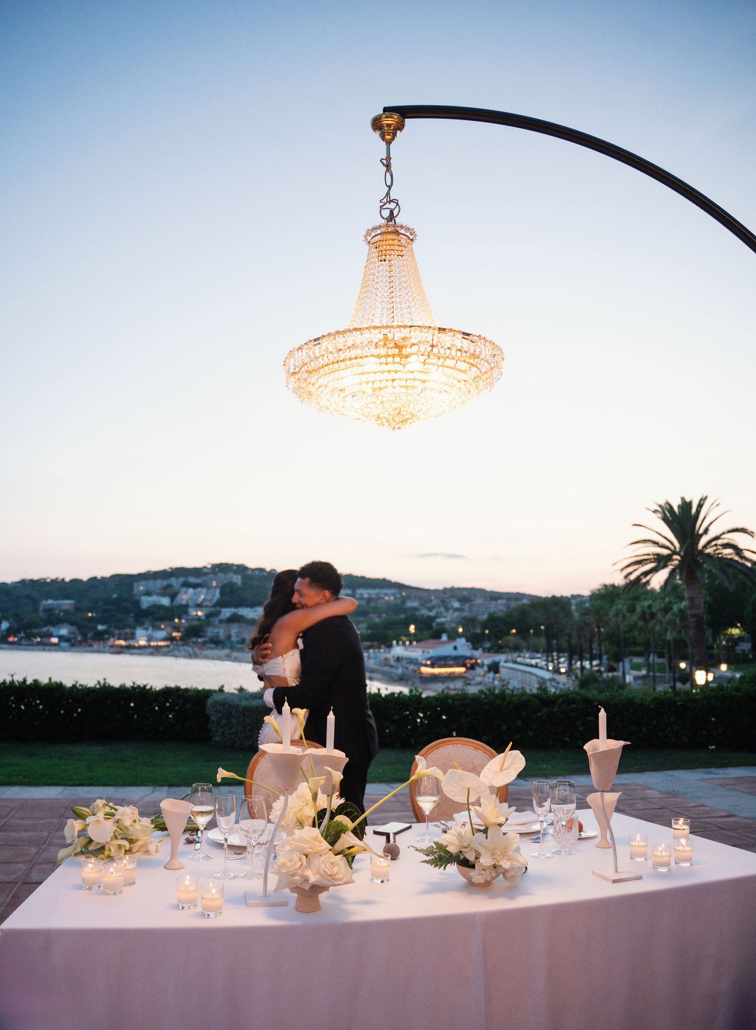 Elegant wedding reception setup with crystal chandeliers and white floral centerpieces along a seaside venue at dusk.