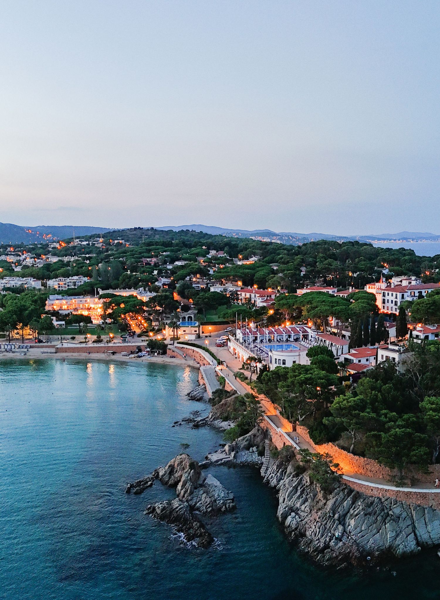 Aerial view of a coastal town with red-roofed houses and winding roads along turquoise Mediterranean waters at sunset.