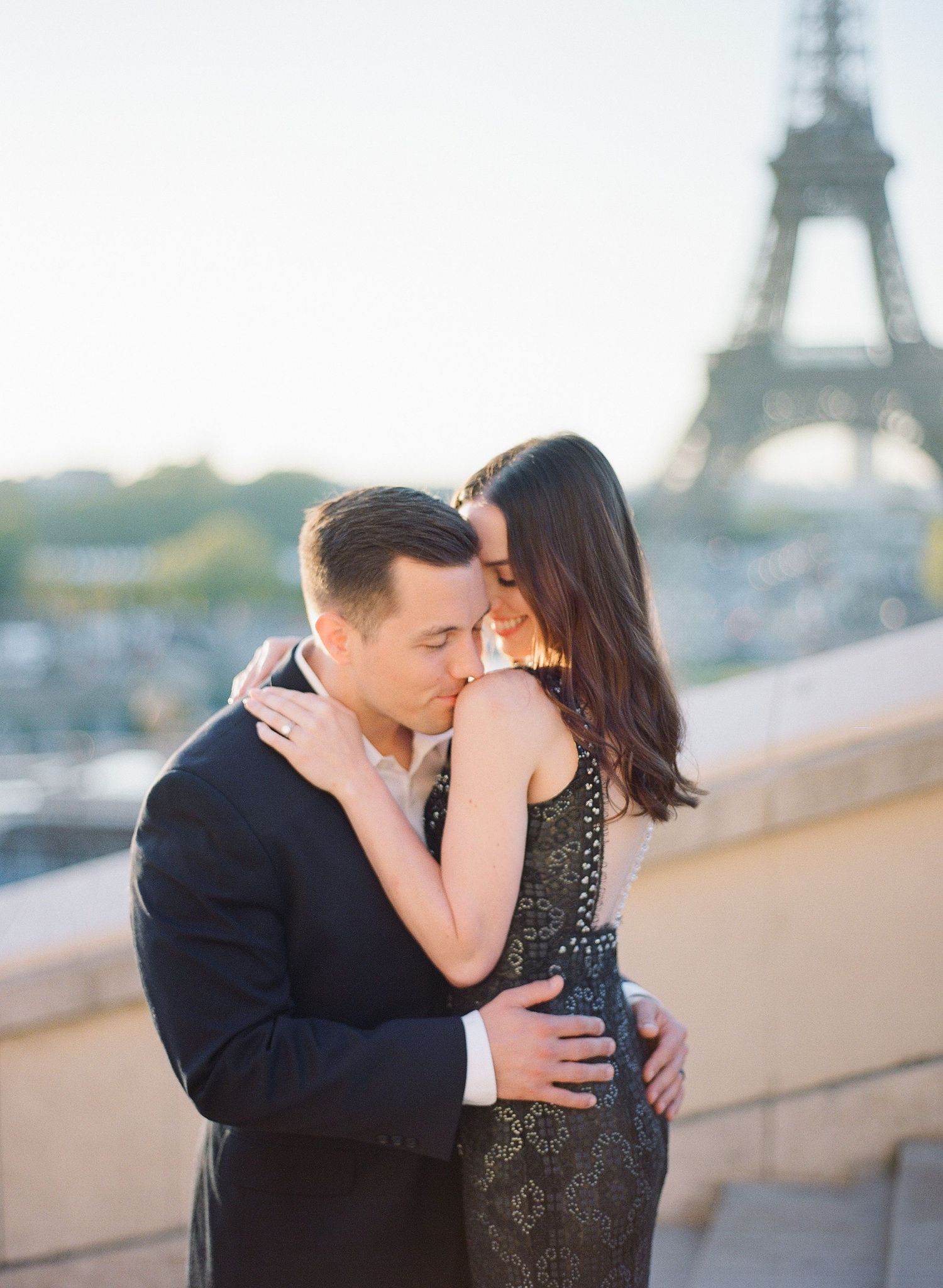A romantic couple shares intimate kisses with the iconic Eiffel Tower in Paris visible in the background during sunset.
