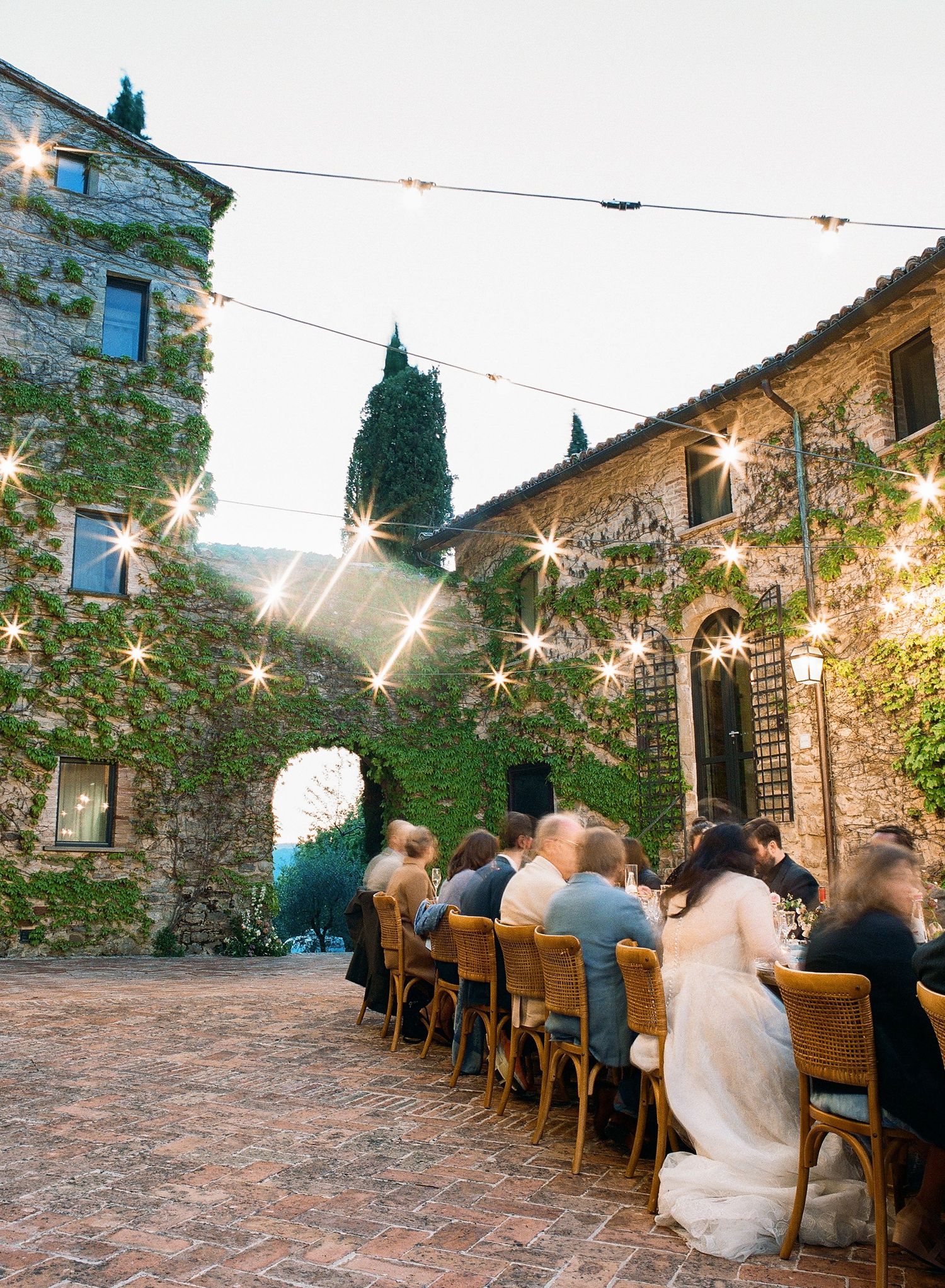 String lights illuminate a rustic stone courtyard patio with wooden tables and chairs set for outdoor dining at dusk.